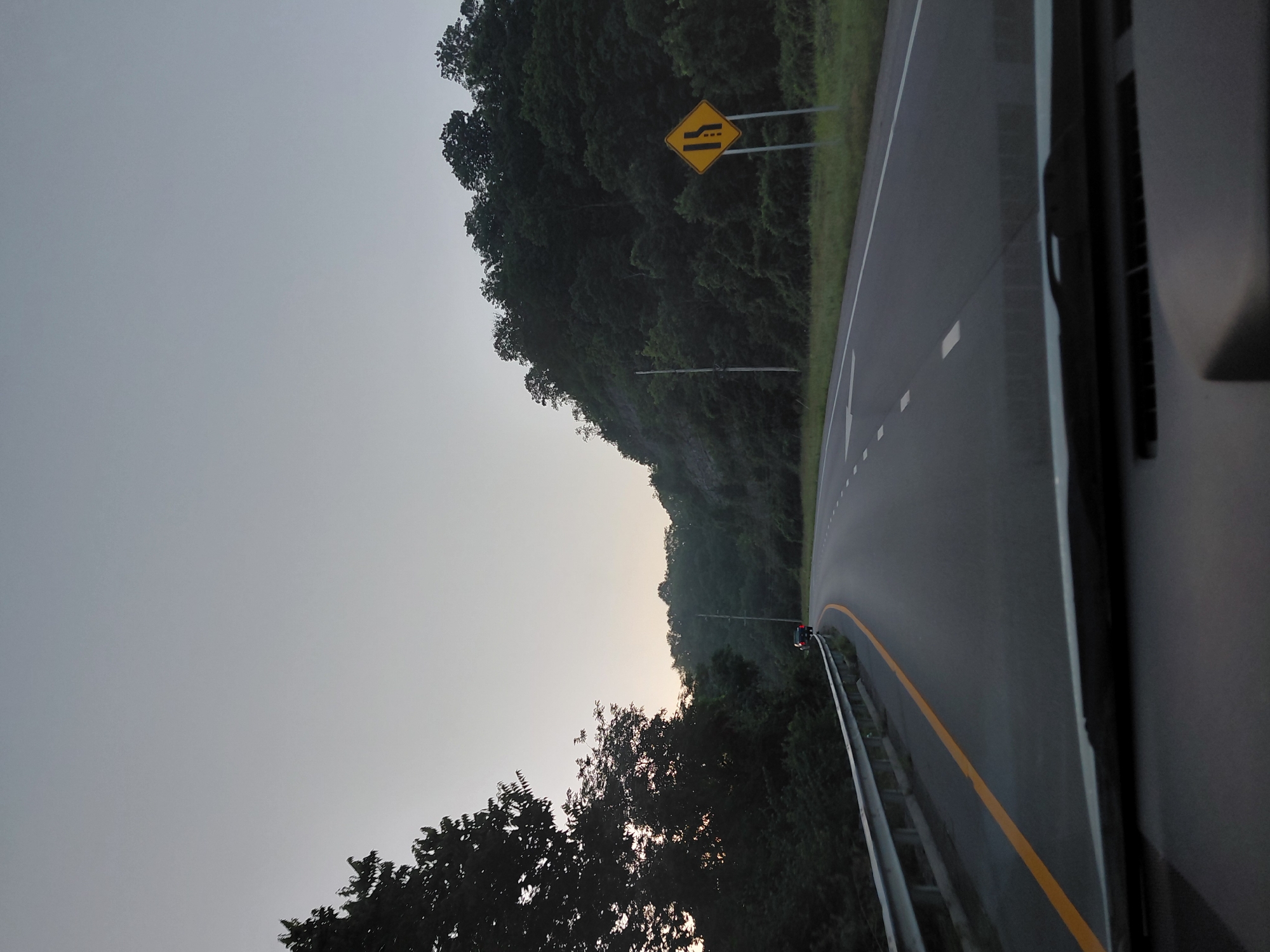 The image captures a view from a vehicle driving on a highway towards a forested hill. The sky is overcast and a pale gray, indicating early morning or late evening. On the left side of the road, a guardrail runs along the shoulder, bordered by a yellow line. Dense trees line the embankment to the left. On the right side of the highway, a yellow diamond-shaped sign with black markings indicates a lane reduction. Beyond the sign, the highway curves around a steep, tree-covered hill. A few cars are visible on the road ahead, some pulled over on the shoulder near the guardrail. The overall scene suggests a rural highway setting.