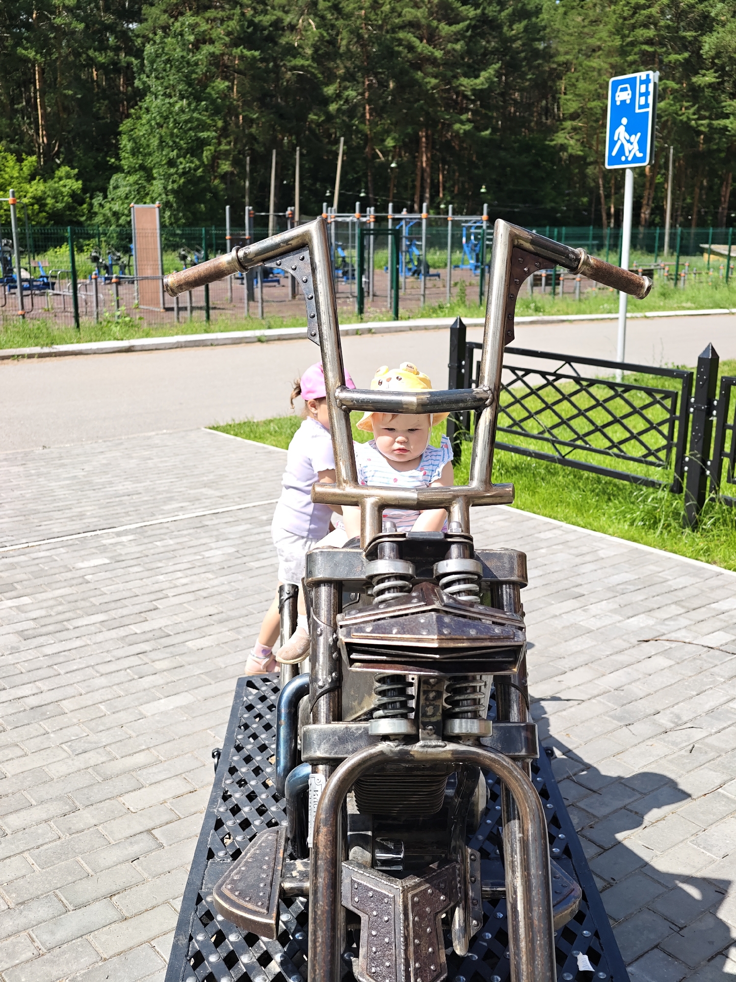In Chaikovsky, Russia, two young children are on a decorative motorcycle, possibly a playground feature, situated on a paved area adjacent to a grassy park. The motorcycle is a replica, constructed from metal with visible springs and decorative elements. One child, a baby wearing a yellow sun hat, is seated in the driver's position, looking forward. Another child, wearing a pink hat and white t-shirt, stands behind the motorcycle, their face obscured. In the background, a fenced-off area with playground equipment and exercise machines is visible, bordered by a dense forest of pine trees. A blue traffic sign indicating a pedestrian crossing is also present. The day is sunny, casting shadows on the pavement.