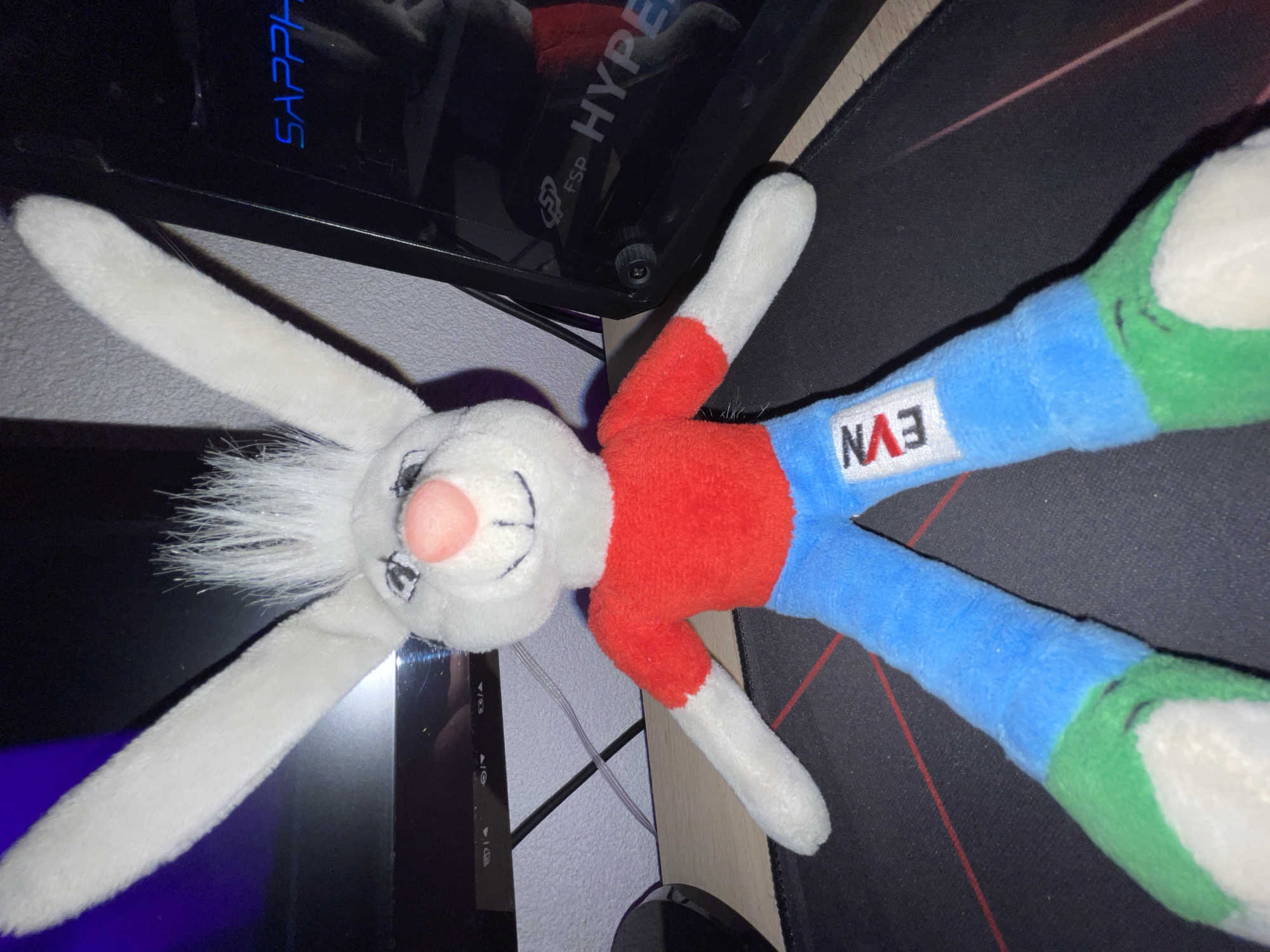 The image displays a white plush rabbit toy lying on a dark surface, likely a desk. The toy is predominantly white, with a pink nose and stitched black eyes and a smiling mouth. It has long, floppy white ears and short, spiky white synthetic hair around its face. The rabbit is dressed in a red short-sleeved top and blue long pants, which terminate in green feet. On the blue pants, the letters "EVN" are visible in white text, oriented upside down relative to the rabbit's head.
The plush toy is positioned diagonally across a black mouse pad, which features subtle red linear patterns. In the upper part of the background, a dark electronic device, likely a computer tower, is visible. On this device, illuminated blue text spells "SAPPHIRE," and other white text shows "FSP HYPER." Cables are also visible near the electronic device. The background further includes a light-colored surface, possibly a wall or wooden desk, suggesting an indoor setting. The scene depicts no activity other than the stationary toy. The lighting appears to be artificial.