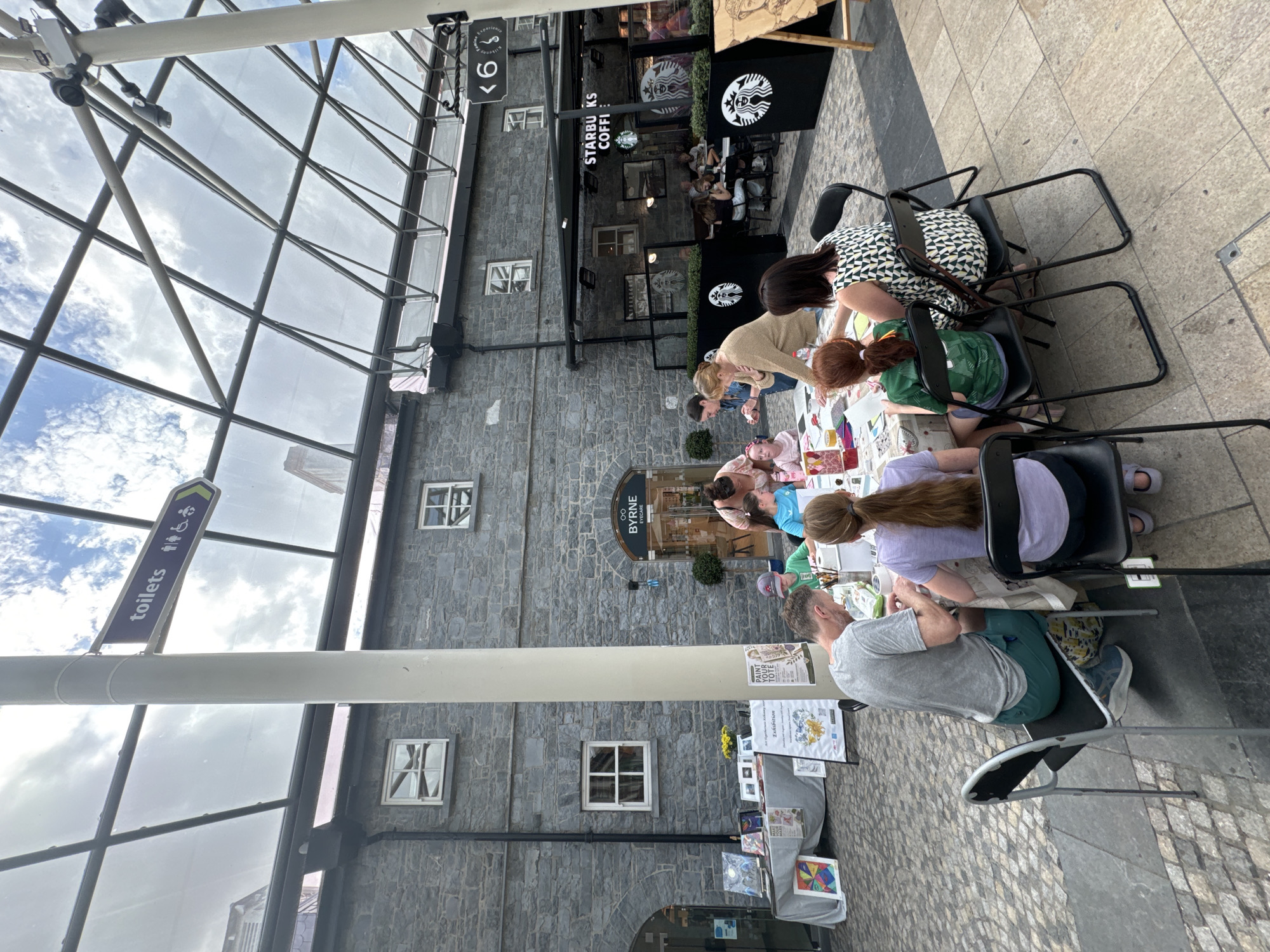 This image depicts an outdoor art workshop for children set against a backdrop of a stone building with arched doorways and windows, characteristic of Kilkenny, Ireland. The workshop is situated under a glass and metal-framed ceiling, possibly part of a modern extension to the historic architecture.
In the foreground, several people are gathered around a table where art supplies are laid out. Two adults, a man and a woman, are seated and appear to be supervising or assisting the children. The man, wearing a grey t-shirt and green shorts, is focused on something on the table. The woman, in a patterned dress, is also engaged with the activity. Two young girls, one with long auburn hair wearing a green t-shirt and another with long blonde hair wearing a purple t-shirt, are seated and actively participating in the art project. Other individuals are partially visible, indicating a larger group is present.
To the left of the workshop area, a display table showcases various artworks, including paintings and prints, suggesting a potential exhibition or sale. A sign that reads "BYRNE EYECARE" is visible on the stone building. In the background, a Starbucks coffee shop is identifiable by its signage and iconic green and white logo, indicating a commercial area within the city. A directional sign pointing to "toilets" with pictograms for men, women, and disabled access is also present, mounted on a light pole that supports the overhead structure. The ground is paved with cobblestones and square tiles. The sky, visible through the glass ceiling, is partly cloudy.
