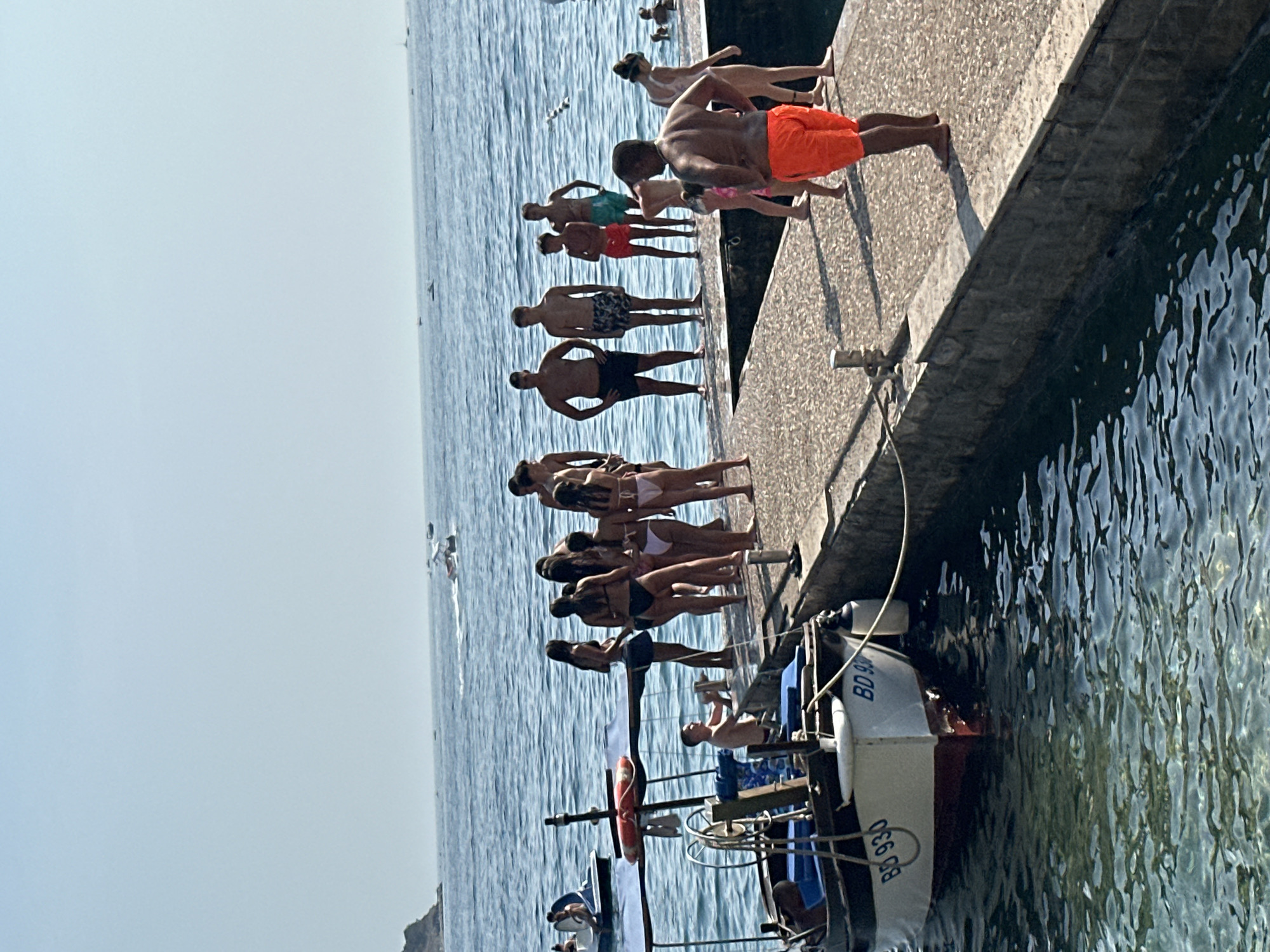 In Budva, Montenegro, a group of people, mostly young adults and teenagers, are gathered on a concrete pier next to a body of water. The day is sunny with a clear blue sky and calm water. Several individuals are wearing swimwear, suggesting a recreational setting like a beach or harbor.
A small motorboat, identified by the markings "BD 930" and "BD 90", is moored to the pier on the left side of the image. The boat has a white hull with red trim and appears to be a fishing or leisure vessel. The pier itself is made of concrete blocks and has metal bollards for mooring.
In the background, the sea stretches out to the horizon, with several other boats visible in the distance, including a jet ski. A small, rocky island or headland is visible on the far left of the frame. The overall scene depicts a lively summer day at the Budva coast, likely a popular spot for swimming and boating.
