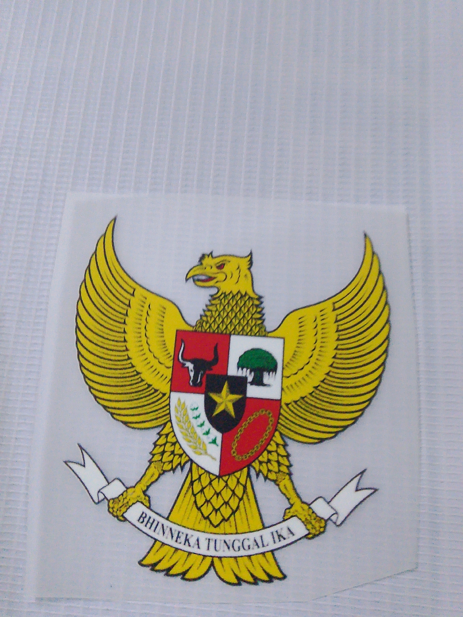 The image displays the coat of arms of Indonesia, Garuda Pancasila, printed on a white, semi-transparent sheet. The Garuda, a mythical bird with golden feathers and outstretched wings, is depicted centrally. Its chest bears a shield divided into four quadrants. The top left quadrant shows a black bull's head on a white background. The top right quadrant features a banyan tree on a white background. The bottom left quadrant contains a gold star on a black background. The bottom right quadrant shows a red and white striped field with a gold chain. Below the shield, a white banner is held in the Garuda's talons, inscribed with the national motto "BHINNEKA TUNGGAL IKA" in black lettering. The background is a textured white surface, possibly fabric, with subtle vertical ribbed lines. There are no people or other objects visible in the image, and no specific location in the city of Semayan, Indonesia, is identifiable.