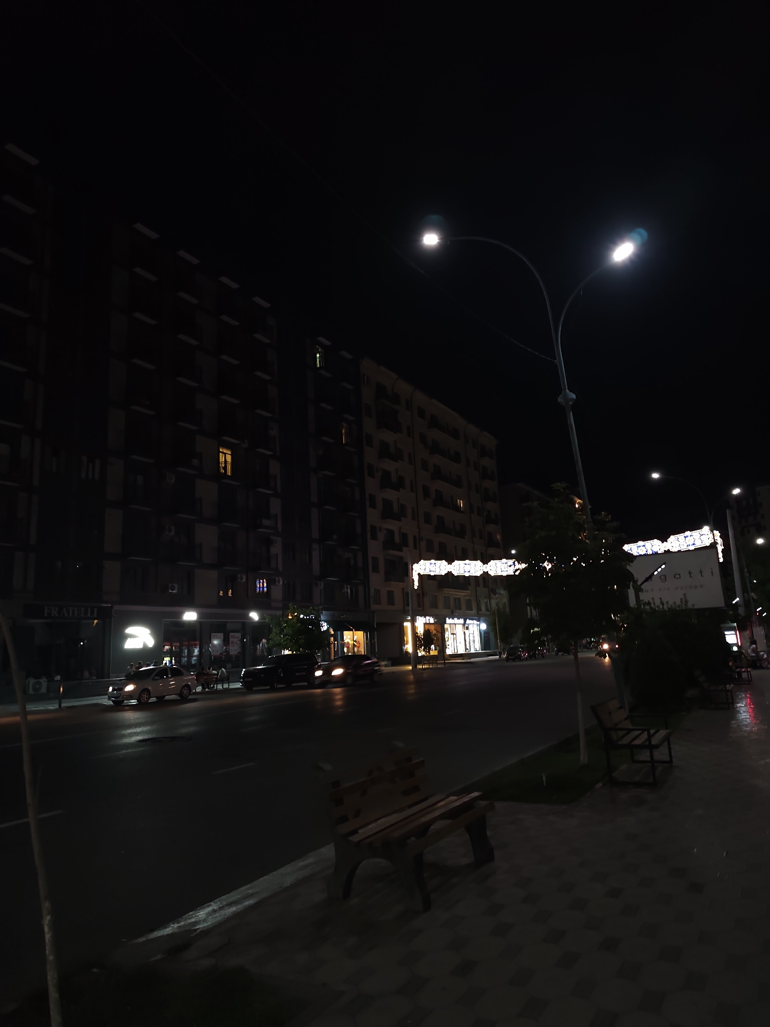 The image depicts a street scene at night in Fergana City, Uzbekistan. A wide, paved street runs horizontally across the frame, with parked cars and streetlights illuminating the asphalt. The street is lined with multi-story buildings, some of which have illuminated windows. On the sidewalk to the right, there are benches, and the ground is paved with interlocking bricks. In the background, a large billboard with illuminated text is visible. The sky is dark, indicating nighttime. There are no people clearly visible in the foreground or mid-ground.