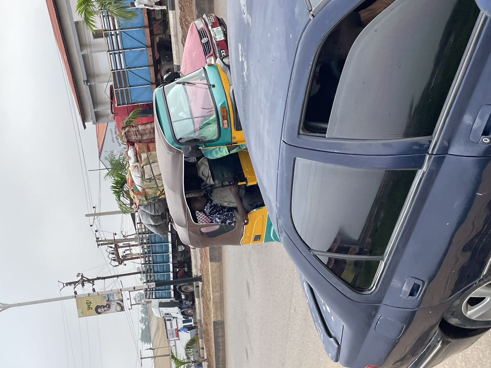The image depicts a street scene in Jos, Nigeria, with a focus on vehicles and infrastructure. In the foreground, the rear section of a dark blue sedan is visible. Behind it, a colorful auto-rickshaw, often called a "Keke Napep" in Nigeria, is prominently featured. This auto-rickshaw is laden with various goods, including sacks, textiles, and what appear to be palm fronds, suggesting it is used for transport. Inside the auto-rickshaw, at least two individuals are visible.
Further back, a blue flatbed truck loaded with more goods, including sacks and wrapped items, is parked or moving slowly. Adjacent to this truck is a red passenger vehicle. In the background, there are buildings with corrugated roofs and a white structure with signage, one of which displays the word "YES!" with an image of a smiling person. Utility poles with numerous wires crisscross the sky, typical of urban infrastructure. The overall atmosphere suggests a busy commercial area in Jos, with vehicles engaged in the transport of goods. The lighting indicates it is daytime, possibly overcast.