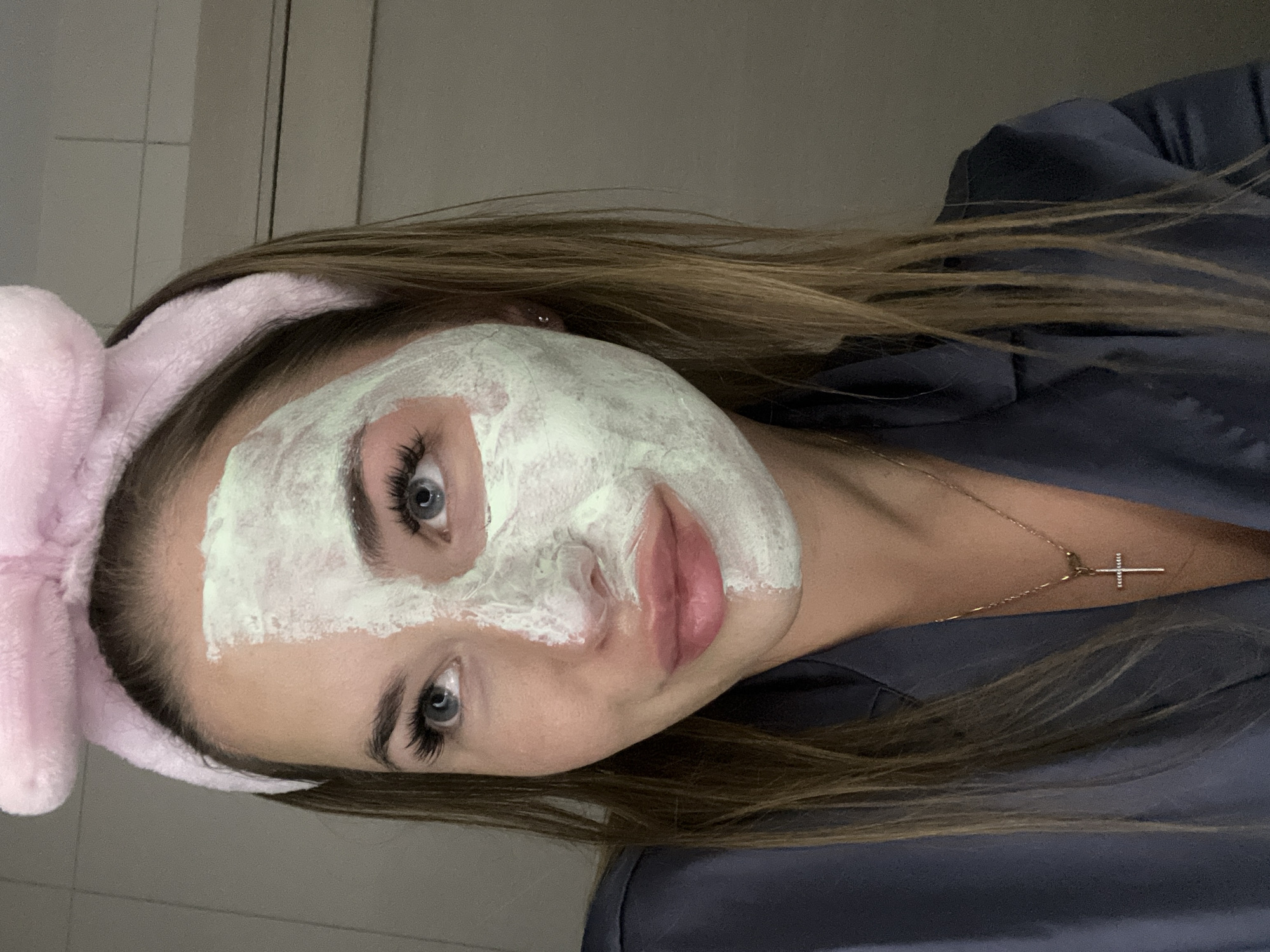 A woman with long brown hair, wearing a pink headband with a bow, has a white face mask applied. She is also wearing a dark blue or grey robe. A delicate gold necklace with a cross pendant is visible on her neck. The background appears to be a bathroom with white tiled walls. The location is not discernible from the image.