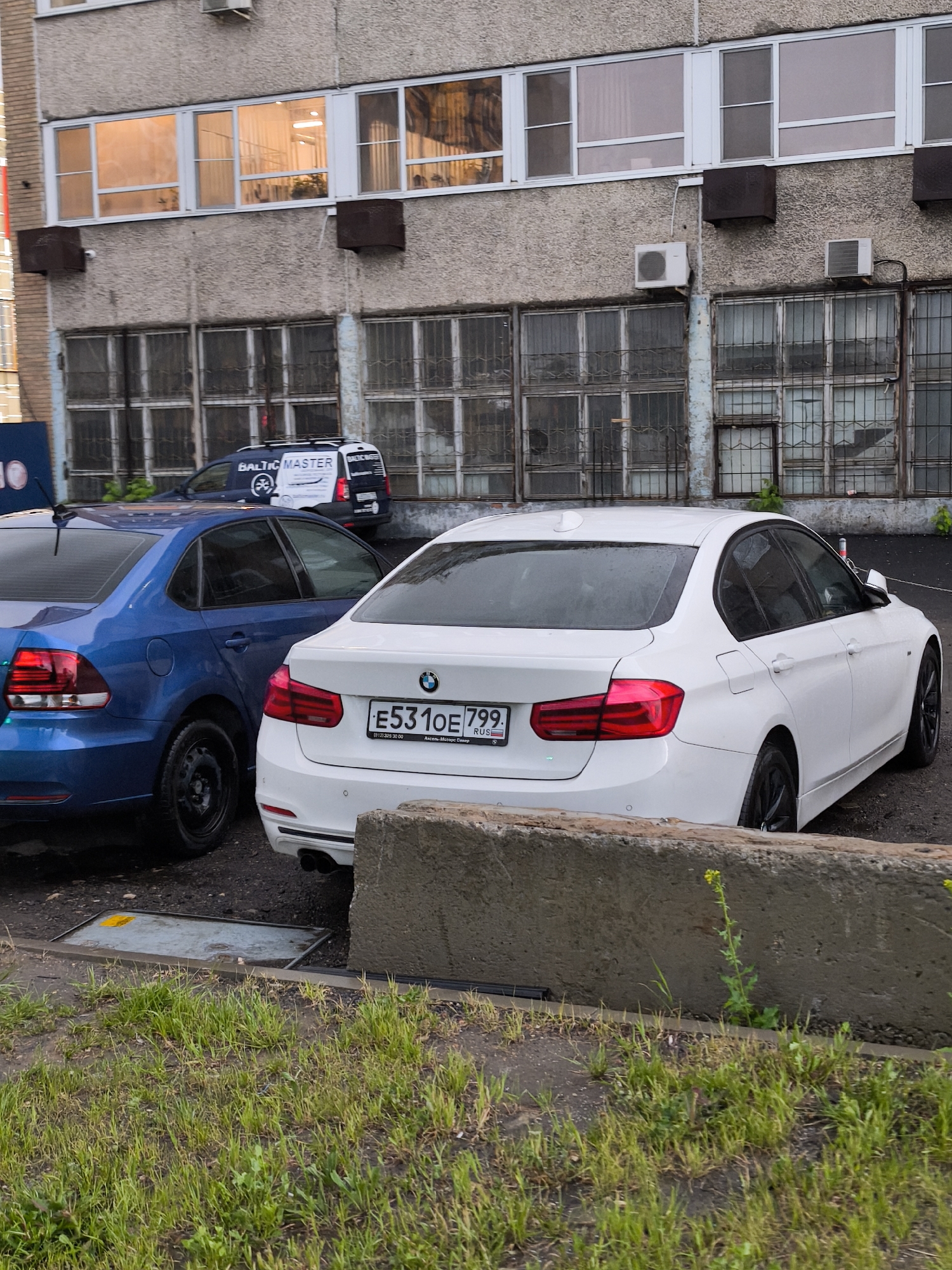 The image depicts a street scene in Moscow, Russia, with three vehicles parked near a concrete building. In the foreground, a white BMW with license plate E531OE799 is parked beside a blue Volkswagen Polo, both facing right. Behind these cars, partially obscured, is a white delivery van with the words "BALTIC MASTER" on its side. The building in the background is made of gray concrete and features several windows, some with bars. The location appears to be a parking area adjacent to the building, with patches of grass and a concrete barrier visible in the lower part of the image. No people are visible in the scene.