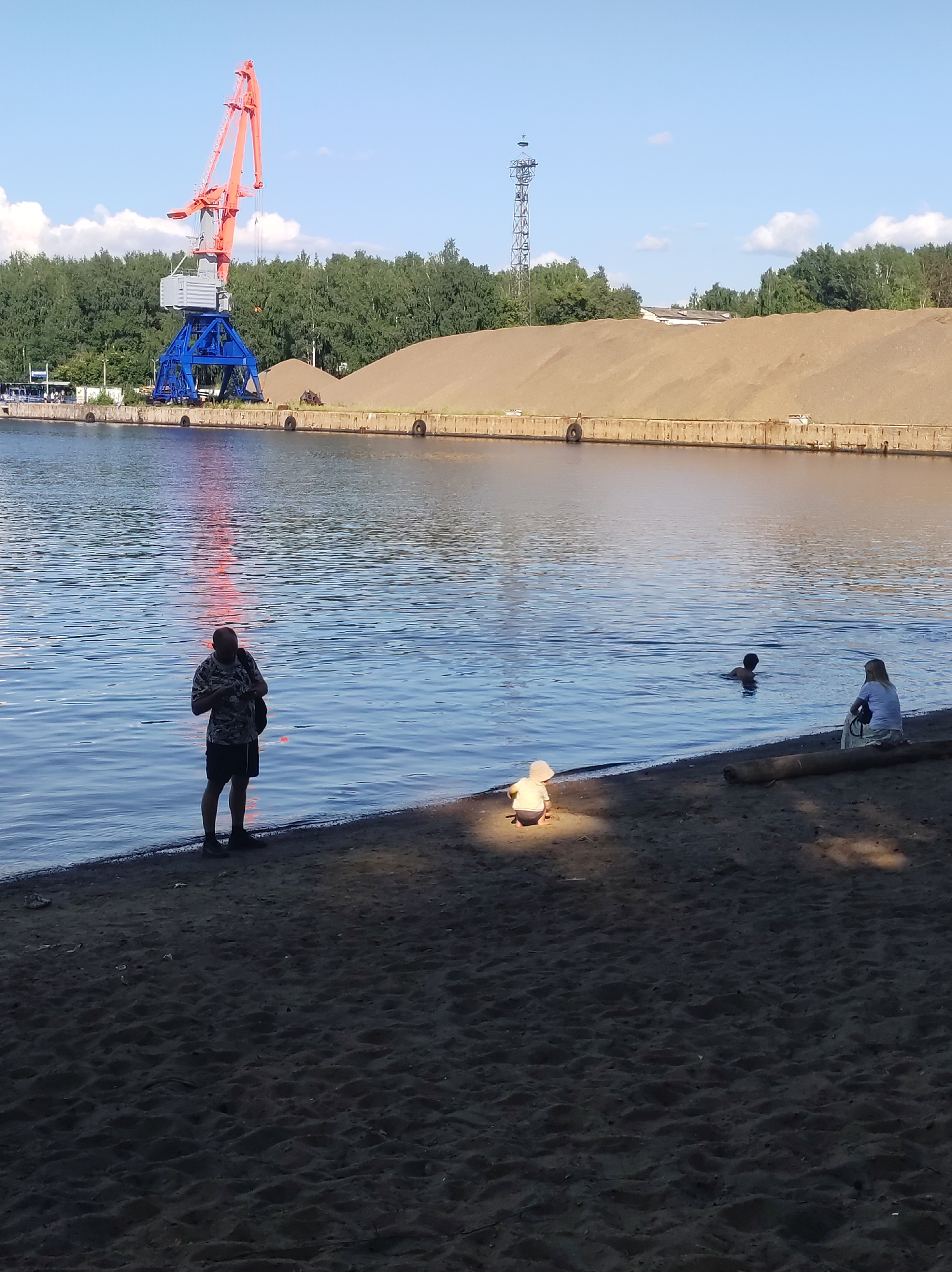 The image depicts a sunny day scene at a waterfront, likely in Chaikovsky, Russia. A large industrial crane, painted orange and blue, dominates the background on the left, situated next to a pile of sand and a concrete embankment lining the water. A tall, metallic tower with antennas stands further in the distance.
In the foreground, on a sandy shore, several people are visible. A man in a camouflage t-shirt and black shorts stands near the water's edge, looking at his phone. Further along the shore, a child in a light-colored hat and shirt is playing on the sand, illuminated by a patch of sunlight. Two other individuals are in the water, one swimming and another sitting on the shore. A fallen tree trunk rests on the sand near the seated person. The water reflects the bright sky and some reddish hues, possibly from the sunset or the crane. The overall atmosphere appears relaxed, with people enjoying a summer day by the water. The weather is clear and sunny.