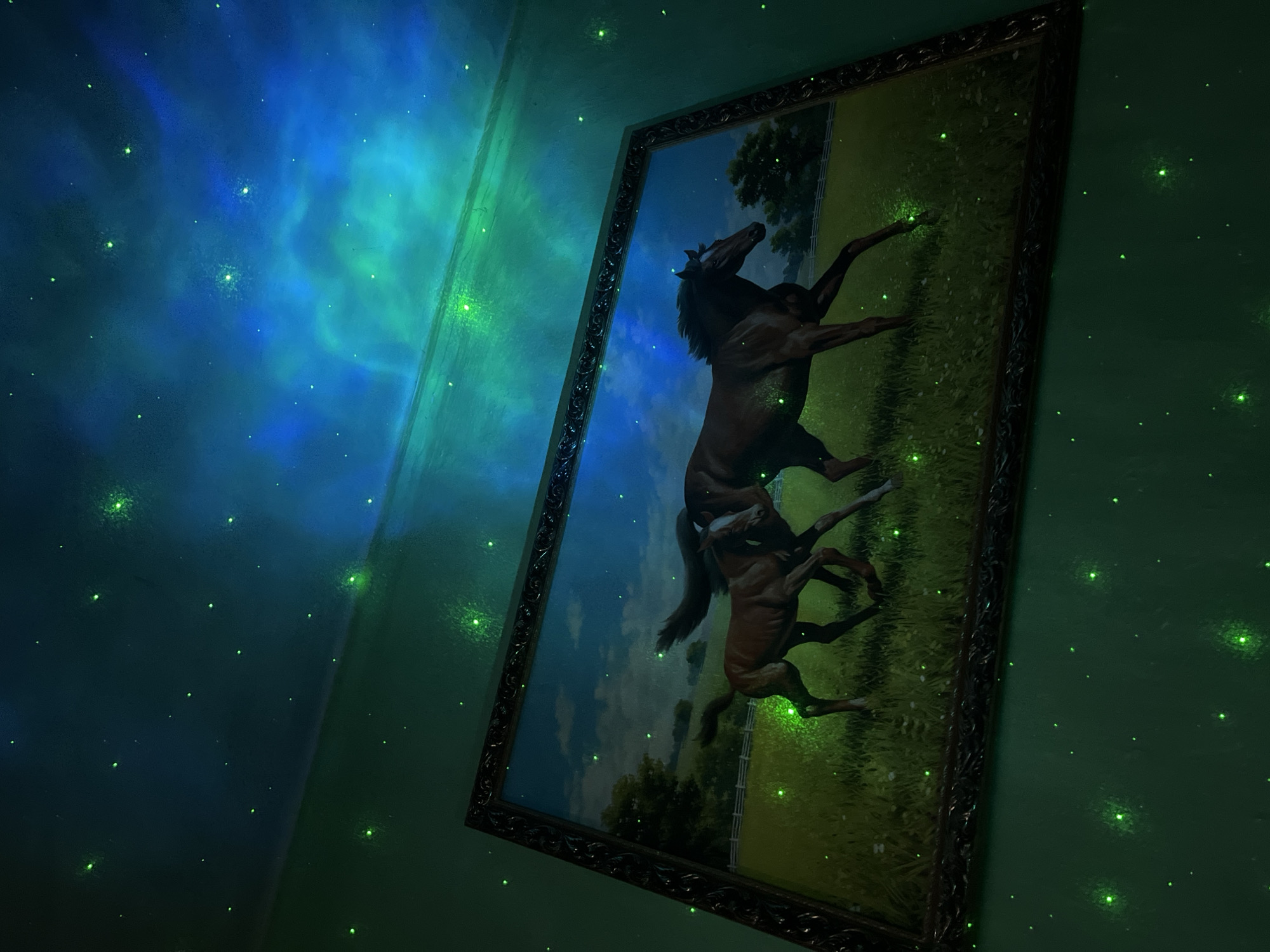 A framed portrait of horses is hung on a wall illuminated with a green-tinted constellation pattern. The portrait, in a dark decorative frame, depicts two brown horses in a field under a blue sky with scattered clouds. The wall itself is a solid color, overlaid with the constellation pattern. The lighting creates a somewhat surreal, almost dreamlike atmosphere within the room. The exact location within Nizhny Tagil, Russia, cannot be determined from the image.