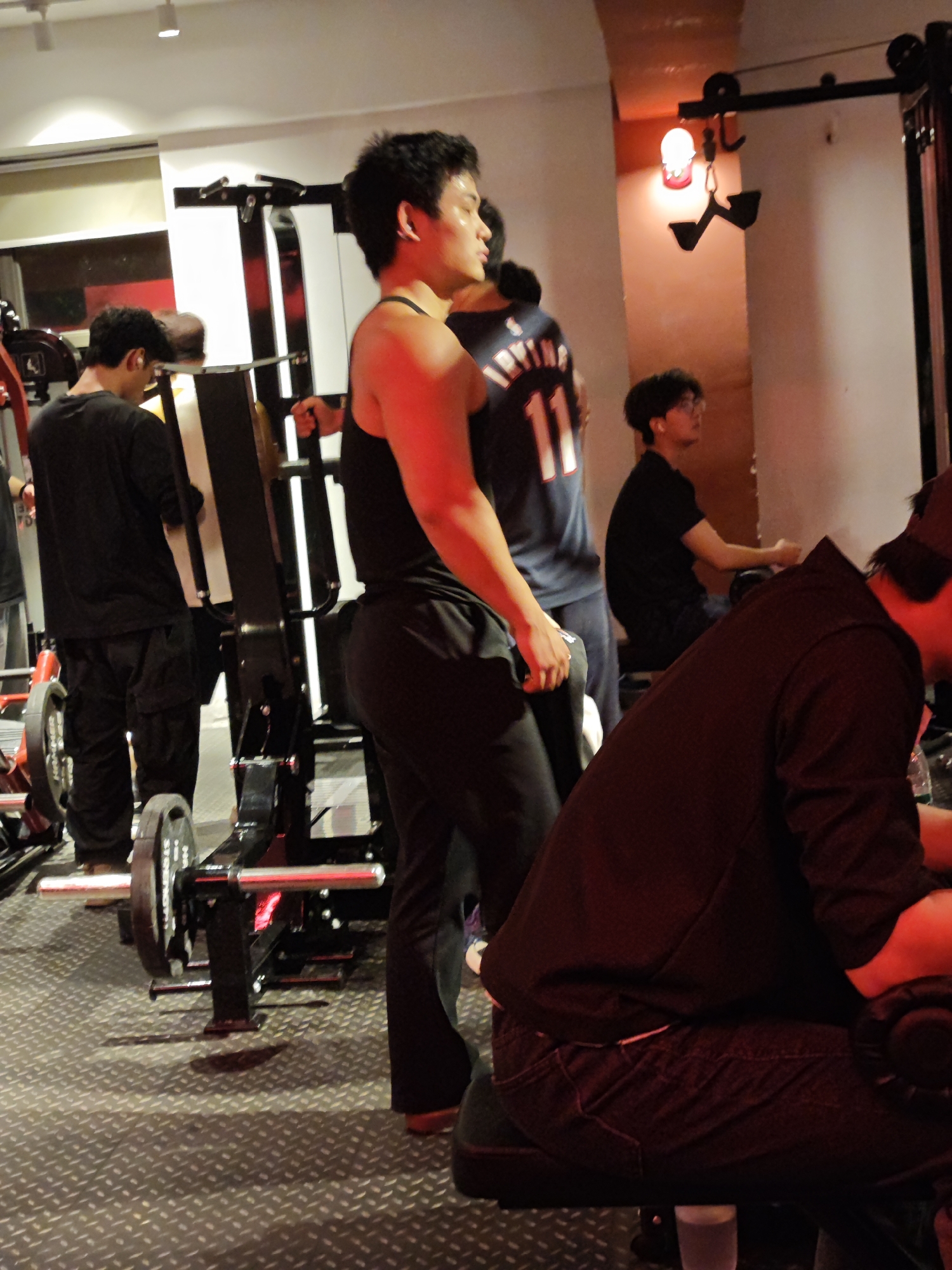 The image depicts a gym scene in the High-tech Zone of Chongqing, China. Several individuals are present, engaging in various activities. One person is positioned near a weightlifting machine, dressed in athletic wear. Another is wearing a basketball jersey, and another is seated. Gym equipment is visible, and the floor has a textured, metallic surface. The lighting is a mix of artificial and natural, with some areas appearing brighter than others.