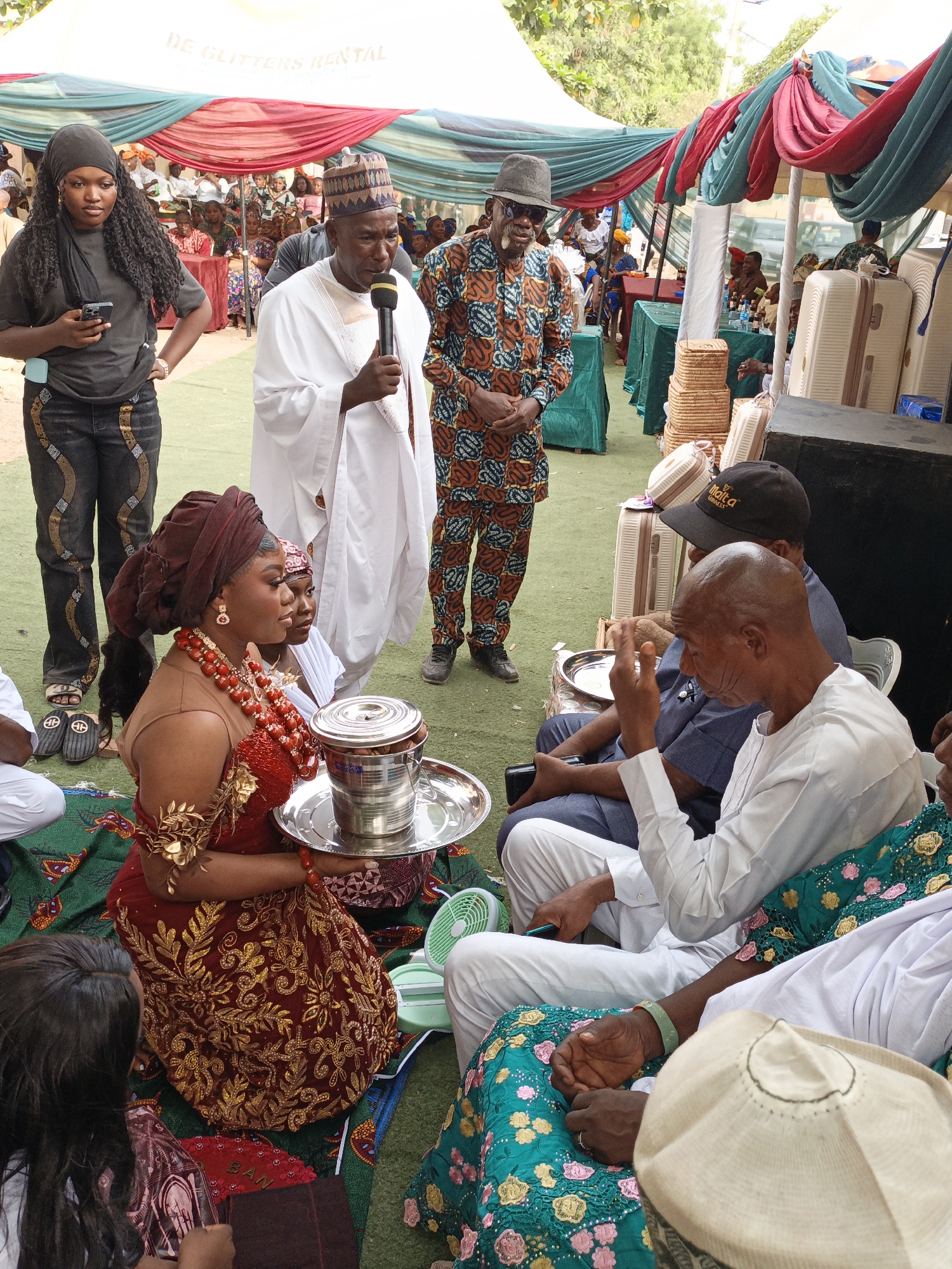 This vibrant, eye-level photograph captures an outdoor ceremony or celebration in Sabon Gari West, Nigeria, likely during the daytime. The event is set under a large tent draped with red, blue-green, and white fabric, with the text "DE GLITTERS RENTAL" visible on its canopy. The ground is covered with green turf, with decorative patterned mats laid out for seating.
In the foreground, a woman, likely a bride or honoree, is seated on a green and red patterned mat. She wears a rich red gown elaborately embroidered with gold patterns, a maroon headwrap, and striking red beaded jewelry, including a prominent necklace and arm adornments. She holds a silver tray carrying a lidded metal container. Opposite her, an older man in a white tunic and trousers is seated, gesturing with his hand as he speaks or participates in a ritual. Next to him, another man wears a black cap bearing the text "Malta Guinness."
Standing centrally, a man in a traditional white agbada and a patterned cap speaks into a microphone, indicating a formal address or blessing. To his right, another man in a patterned ankara suit and a dark hat observes the proceedings. On the far left, a young woman with long, dark, curly hair stands, looking at her mobile phone.
The scene includes cultural objects such as stacks of light-colored suitcases and woven baskets, possibly gifts, placed to the right. A small green electric fan rests on the ground. In the background, numerous attendees are visible, seated at tables with drinks, suggesting a significant social gathering. The partially visible text "BAN" is on a red fan-like object on the ground. The overall atmosphere appears festive and engaged.