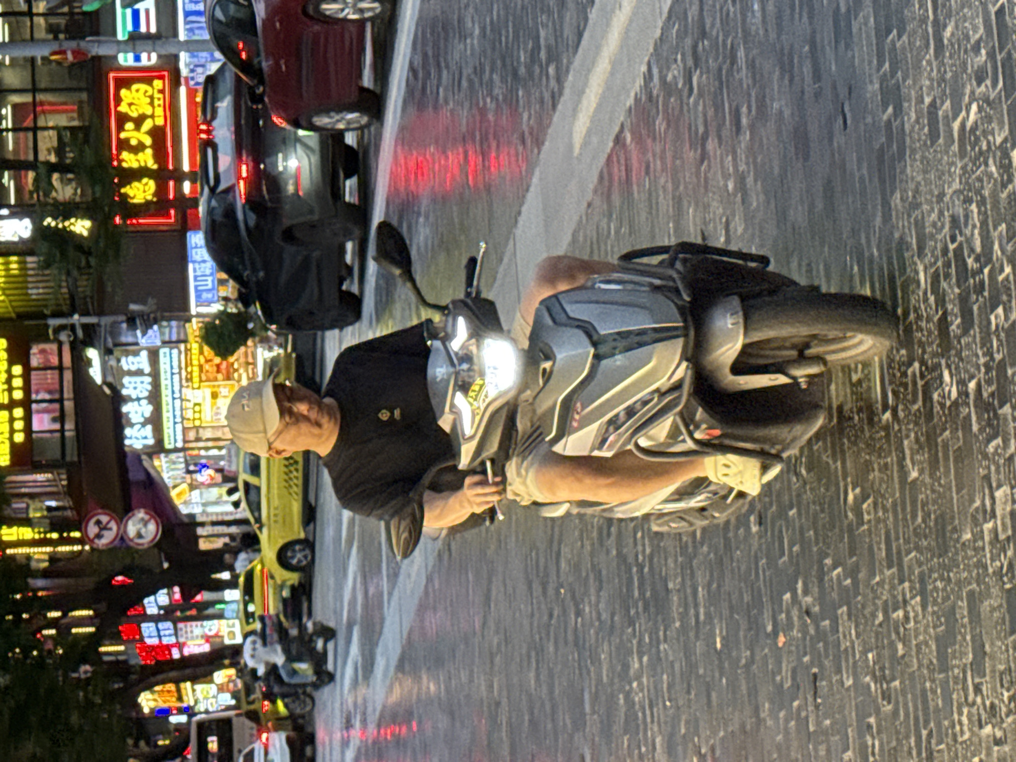 The image depicts a street scene in Yuzhong District, China at night. A man wearing a black t-shirt and tan shorts is riding a silver and gray scooter on a wet, brick-paved road. He is wearing a beige baseball cap and glasses. The scooter's headlight is illuminated. In the background, there are buildings with numerous neon signs displaying Chinese characters in red, yellow, and white. Several vehicles, including a yellow taxi, a black car, and a red car, are visible on the road. Reflections of the neon lights are visible on the wet surface of the street. The overall atmosphere suggests a busy urban environment illuminated by artificial lights.