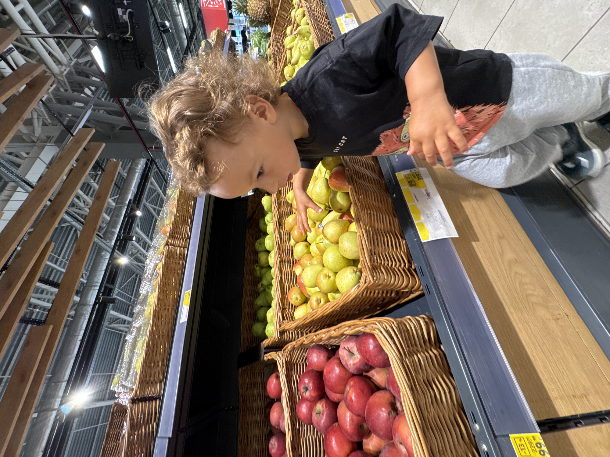 In a brightly lit supermarket, a young child with curly blonde hair stands in front of a display of produce. The child, wearing a black t-shirt with a graphic print and grey sweatpants, reaches out towards a basket filled with green pears. To the left, another basket overflows with red apples. The display shelves are made of wood and grey metal, and the ceiling features exposed industrial beams and lighting fixtures. The overall environment suggests a modern retail setting, likely in a city like Sochi, Russia, given the context. The child's focus is directed at the fruit, indicating an act of selection or exploration.