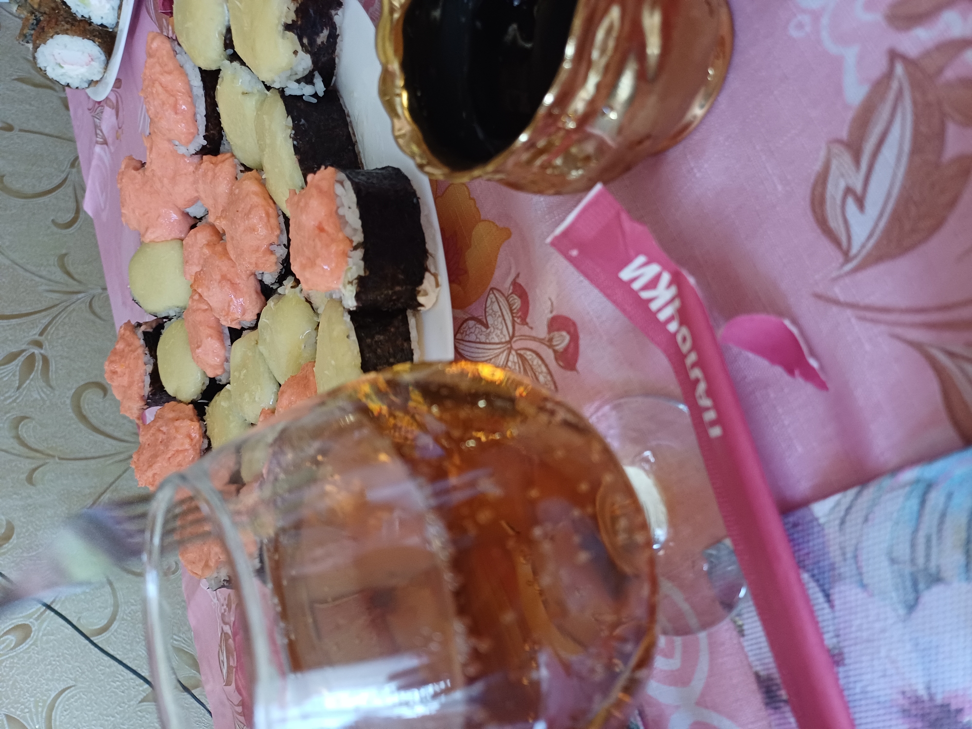 A partially consumed meal of sushi and a beverage is laid out on a pink tablecloth. In the foreground, a glass of amber-colored liquid with visible bubbles sits to the left. In the center, a plate of sushi rolls is arranged, some topped with a creamy pink mixture and others with a pale yellow topping. To the right, a small, ornate golden bowl filled with a dark liquid, presumably soy sauce, is positioned. A pink paper wrapper with Cyrillic lettering that reads "ПАЛОЧКИ" (meaning chopsticks) is also visible. In the background, a floral patterned wallpaper adorns the wall. There are no people visible in the frame.