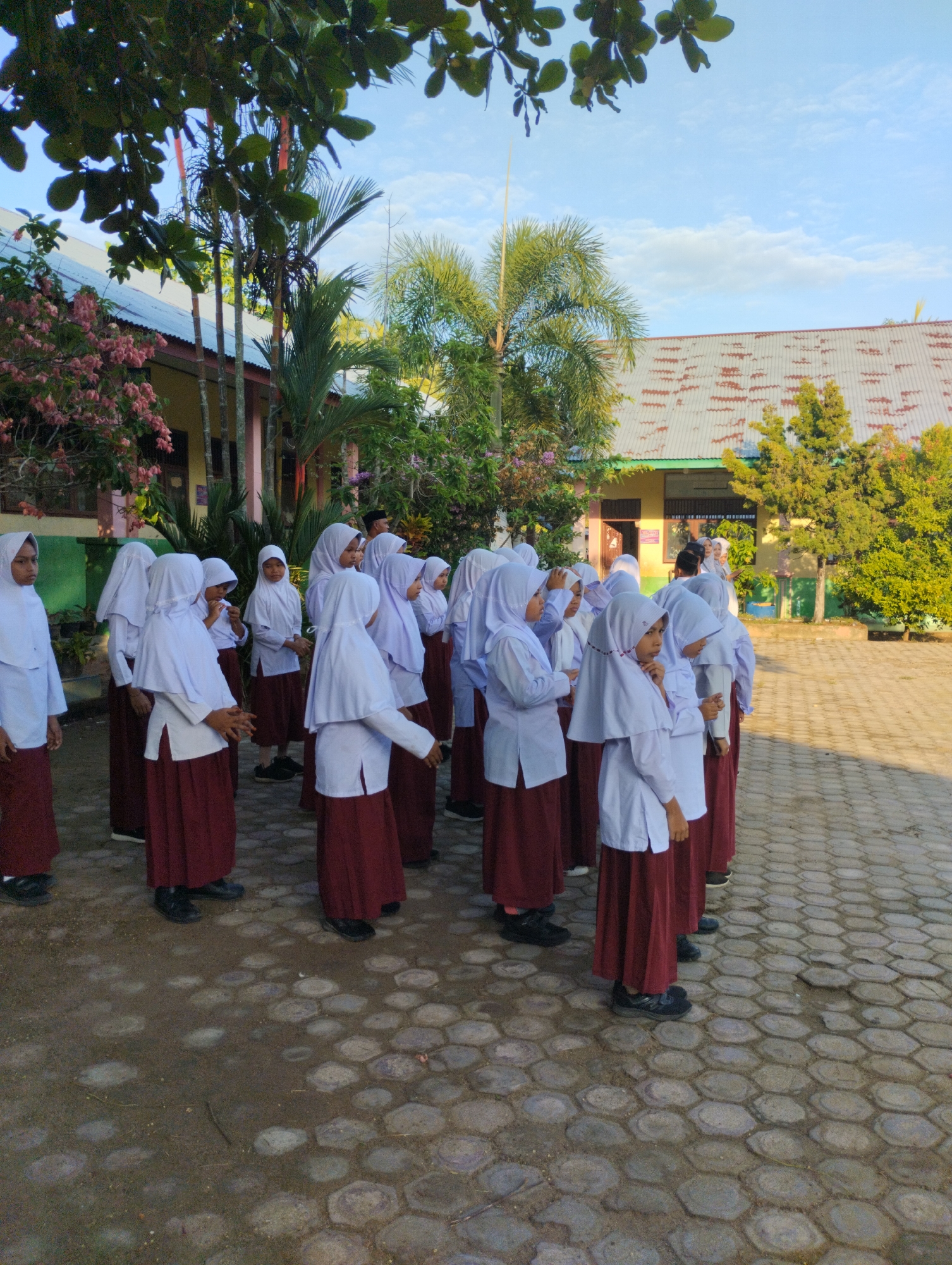 A group of young female students, dressed in white hijabs and maroon skirts, stand in a line on a paved outdoor area. The students appear to be at a school, with buildings visible in the background. The buildings have a light-colored facade with some green trim and corrugated metal roofs. Trees and other vegetation are present around the buildings, suggesting a tropical or subtropical environment. The sky is clear and blue with a few scattered clouds. The pavement is made of interlocking hexagonal pavers. The scene is likely in Baratpayaitek, Indonesia, given the architectural style and vegetation.