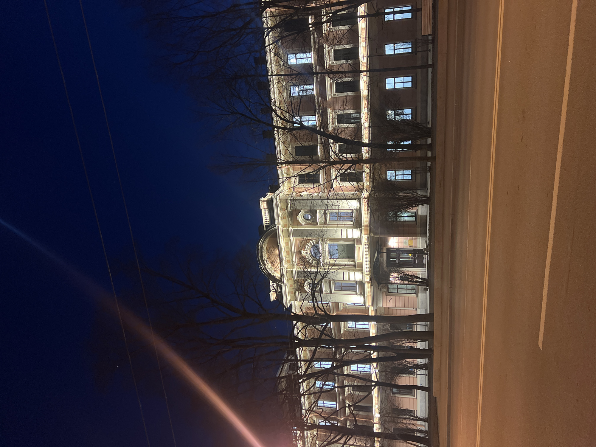This nighttime image depicts the Yusupov Palace at Moika Embankment, Saint Petersburg, Russia. The multi-story building, characterized by its ornate Neoclassical architecture with a light pinkish facade and cream-colored decorative elements, is extensively illuminated by warm architectural lighting. Numerous windows display cool blue-white internal illumination. Bare deciduous trees and dense shrubs stand between the building and the street, partially obscuring the lower levels of the palace. The foreground consists of an asphalt road with a double solid yellow line and a dashed white line, reflecting ambient light. The sky above is dark navy blue, featuring a diagonal light streak, likely a lens flare. No people or vehicles are visible in the scene.