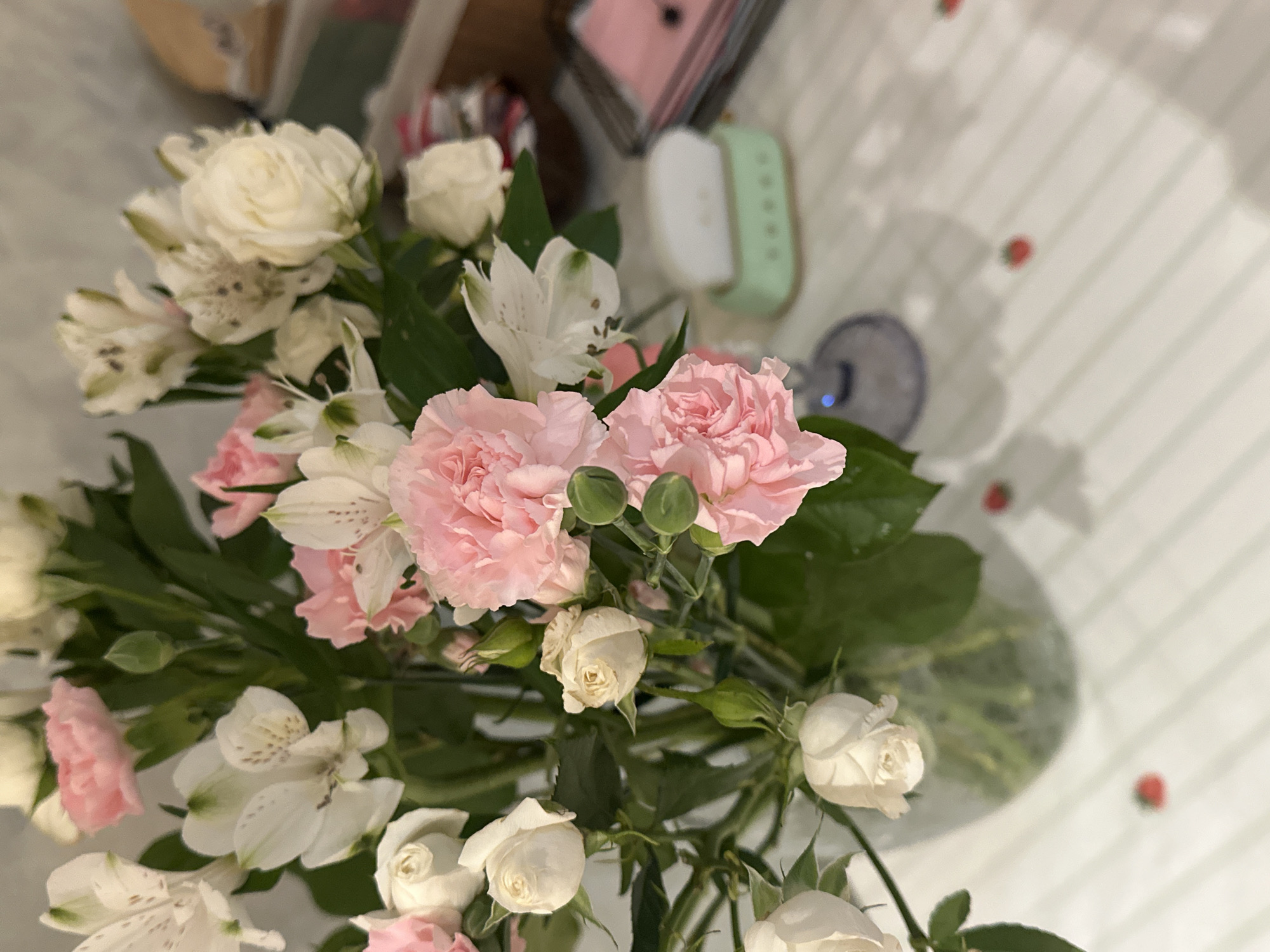 The image is a high-angle shot of a floral arrangement on a white surface with red dots. The arrangement contains pink carnations, white alstroemeria, and small white roses. The flowers are arranged in a vase. The background includes blurry objects such as a smartwatch and other items on a table. The location, Šušanj, Montenegro, is not evident from the image.