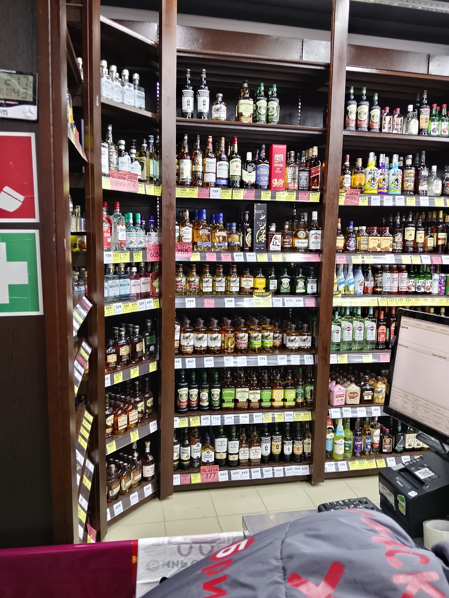 This image depicts a section of a liquor store in Oktyabrskiy, Russia, likely within the city limits. The scene is dominated by tall, dark wooden shelves filled with various bottles of alcohol. The shelves are organized into multiple tiers, showcasing a wide assortment of spirits, including whiskey, vodka, gin, and liqueurs. Many bottles are distinct in shape and color, with labels visible on some. Price tags and sale indicators are present on many shelves, suggesting a retail environment.
In the foreground, a portion of a checkout counter is visible, with a computer monitor and a cash register. A keyboard is also partially visible. The floor is tiled. The overall lighting suggests an indoor setting, possibly under artificial lights common in commercial spaces. There are no people clearly visible in the frame, but the presence of the checkout equipment implies recent or ongoing customer activity. The overall impression is that of a well-stocked retail outlet focused on alcoholic beverages.