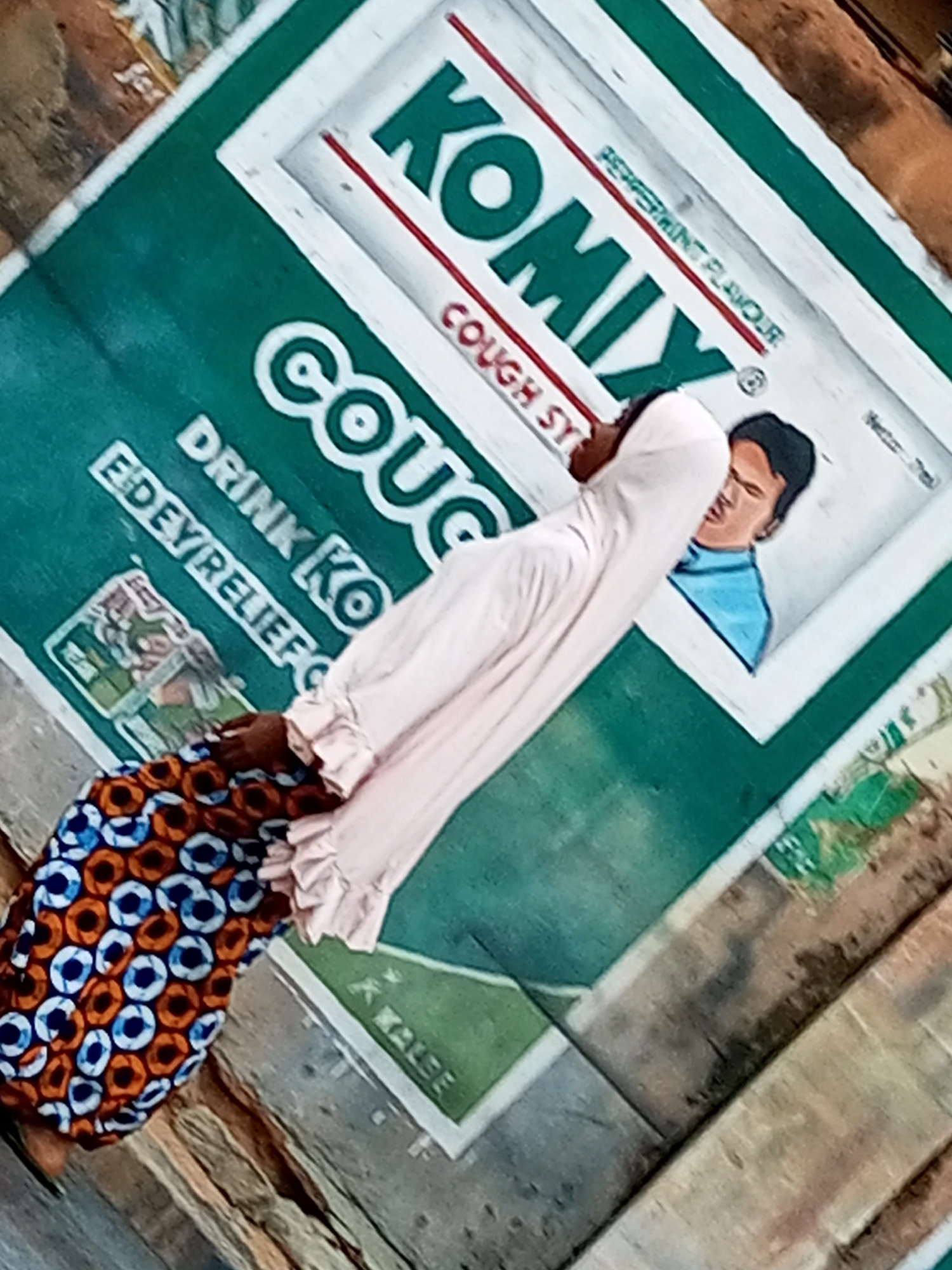 The image shows a person in traditional clothing, with a patterned skirt and a light pink, ruffled top, positioned in front of a large green advertisement. The advertisement features the brand "KOMIX" in large green letters with a red outline. Below this, "COUGH SYRUP" is visible. Text in a white box reads "PEPPERMINT FLAVOUR". There is a drawing of a man's face with dark hair and a blue shirt on the right side of the advertisement. To the left of the person, partially obscured text in white says "DRINK KO" and "EDEYRELIEF". The advertisement appears to be mounted on a weathered wooden surface, suggesting it might be displayed outdoors or in a public space. The overall scene is likely in a market or street setting. The presence of the "KOMIX" brand, a known cough syrup in Nigeria, and the context of the patterned fabric worn by the individual, point towards a location within Nigeria, possibly Gombe, Nigeria, given the prompt's context. The lighting and angle of the photograph indicate it was taken during daylight.