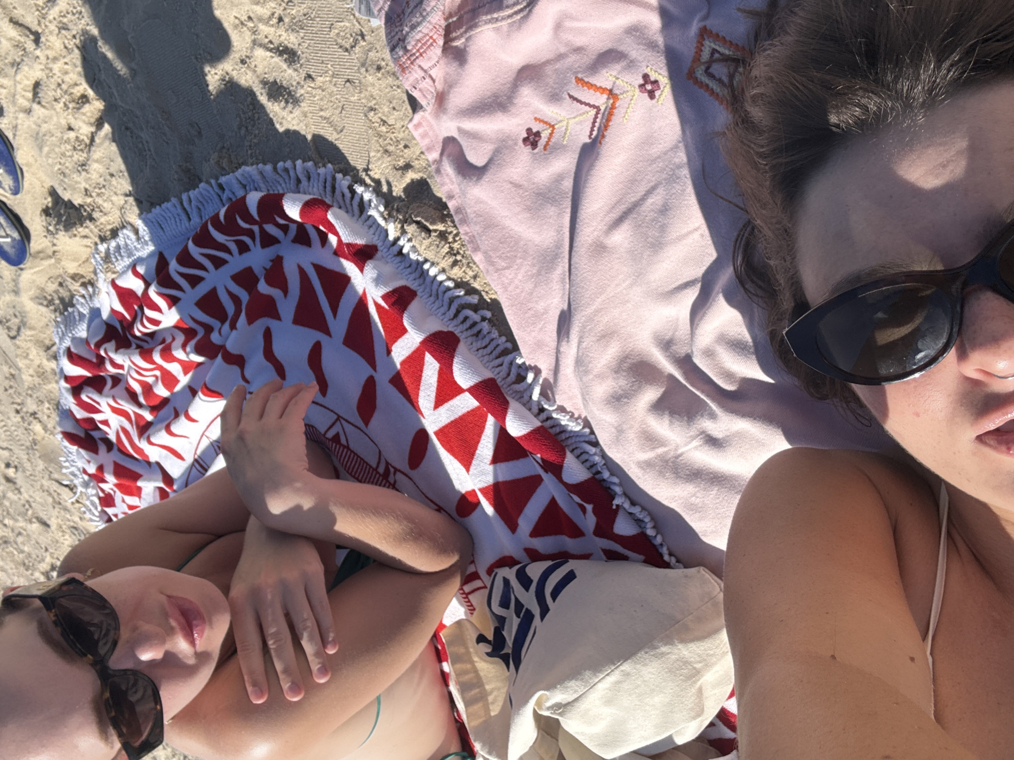 This is a high-angle shot of two people lying on a beach in Tel Aviv, Israel. The scene is set on a sandy beach, indicated by the grainy texture of the ground and the presence of beach towels.
The subjects are two individuals, partially visible from the shoulders up, lying on their backs. The person on the left is wearing sunglasses and a bikini top. Their head is turned to the side, and their arms are crossed. The person on the right is also wearing sunglasses and a bikini top. Their head is tilted back, and their hair is visible. Both individuals appear relaxed and are enjoying a sunny day at the beach.
Several beach towels are spread out on the sand. One is a round towel with a red and white geometric pattern and a fringed edge. Another appears to be a light pink towel with embroidered floral details. A woven tote bag is also visible, with blue and white stripes. The sand shows some footprints and the shadow of what might be a person or an umbrella, suggesting it is daytime. The bright sunlight and the presence of beach accessories strongly indicate a warm, sunny day at the beach.
There is no visible text in the image. The overall mood is one of leisure and enjoyment of a summer day at the beach. The location context of Tel Aviv, Israel, suggests a coastal Mediterranean setting.
