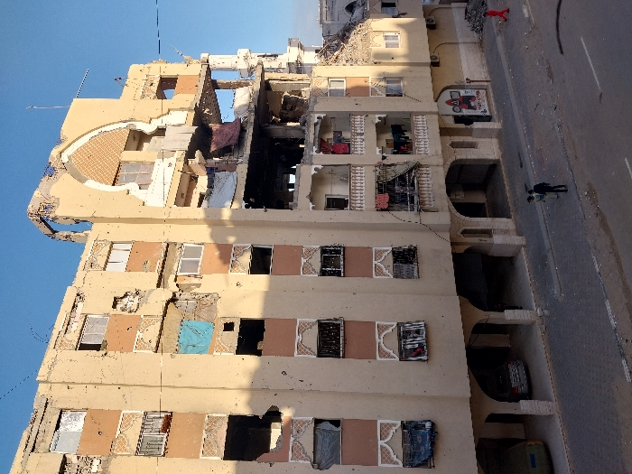 The image displays a multi-story building that has sustained significant damage, standing alongside a paved road under a clear blue sky. The building, constructed with a light beige or tan facade and adorned with some brown or terracotta-colored accents, shows widespread structural compromise.
The upper floors of the building exhibit the most severe damage, with large sections of walls and ceilings completely missing, exposing the interior structure, rubble, and twisted metal. The roofline also appears to be heavily damaged or partially collapsed in parts. Throughout the building's facade, numerous windows are broken or entirely absent, with some still retaining ornate, possibly wrought-iron, grilles. On several balconies and in exposed interior spaces, items resembling laundry or fabric are seen hanging, including red and dark clothes on one upper balcony and a piece of blue fabric on a lower floor. The exterior walls also show signs of impact, such as numerous smaller holes or pockmarks that could be from shrapnel or bullets.
The ground level of the building features a series of arched openings, which could be entrances to residential units, businesses, or parking areas. Within these arched spaces, at least two vehicles are partially visible; a dark-colored sedan is parked in one, and another vehicle is further inside an adjacent archway. On one of the walls at this ground level, a piece of graffiti or a mural is visible, featuring a stylized face or character with red and white elements, though specific text is not discernible.
Adjacent to the building is a paved road, appearing to be a two-lane street, with a curb separating it from the building. Scattered debris, such as small stones and dirt, is visible on the road surface. Two individuals are present on the road or sidewalk: one person dressed in red clothing is walking further down the street, and another person in dark attire is walking closer to the building on the sidewalk.
The lighting suggests it is daytime, with long shadows cast from the building onto the street, indicating either morning or late afternoon. The overall scene conveys a powerful impression of destruction and urban decay, likely the aftermath of a significant event or conflict, with a sense of lingering abandonment despite the presence of people on the street.