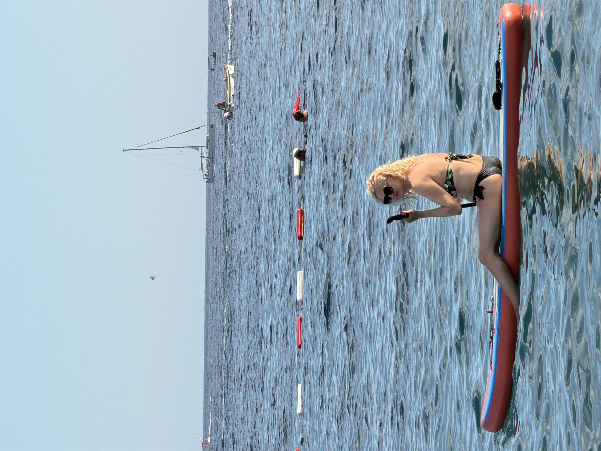 In Budva, Montenegro, a woman with blonde curly hair and sunglasses, wearing a black bikini with a floral pattern, is paddleboarding on a calm, blue sea. She is holding a paddle and appears to be in motion. The sun is shining brightly, casting reflections on the water, indicating it's likely daytime. The background shows the vast expanse of the sea with a clear sky, suggesting good weather. There are other watercraft visible in the distance, including sailboats and what appears to be a paraglider in the sky, hinting at a recreational or tourist area. The overall scene is one of leisure and outdoor activity on a sunny day.