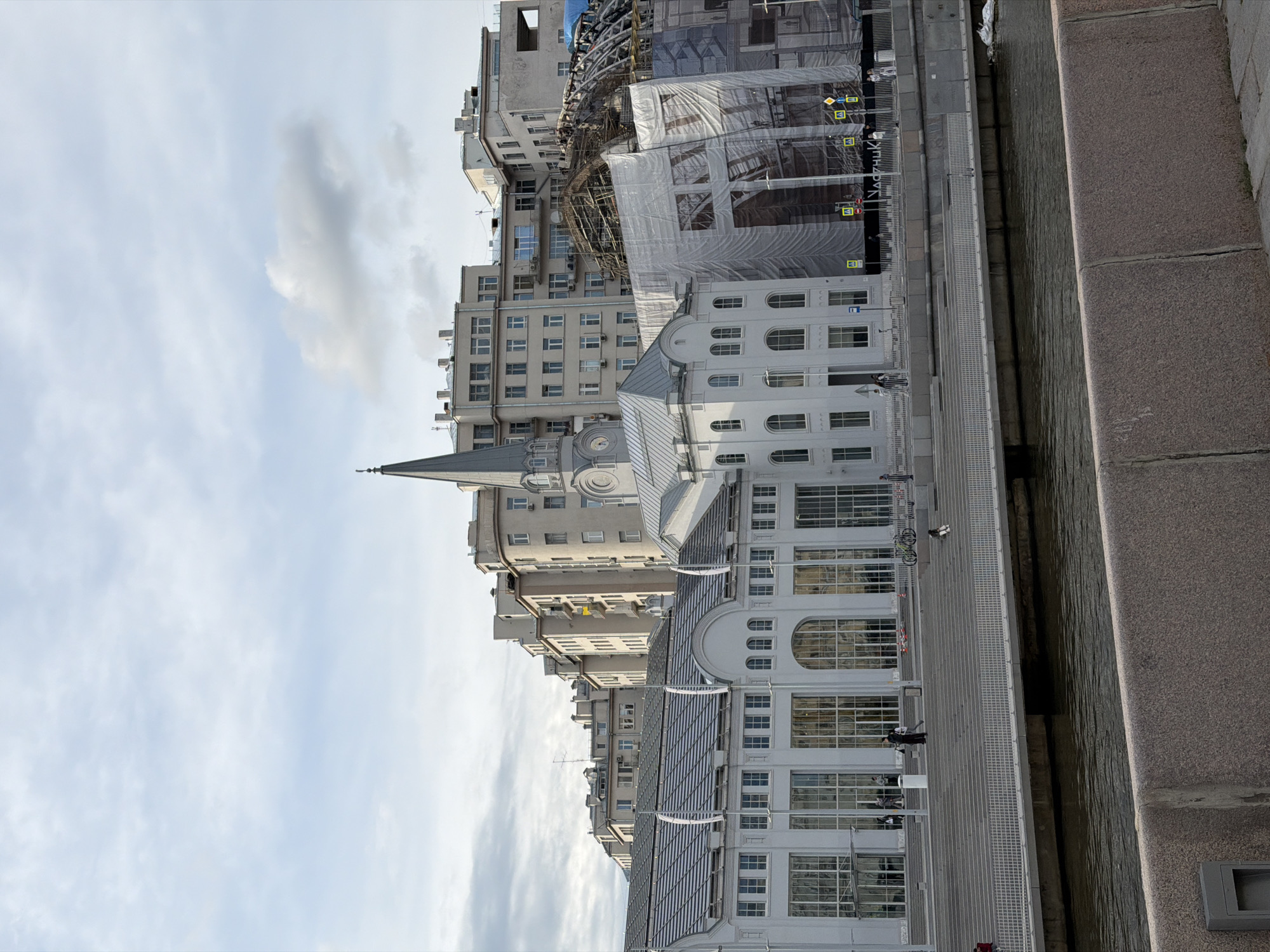The image shows a cityscape in Moscow, Russia, featuring a variety of buildings. There are tall, multi-story buildings with numerous windows, some under construction with scaffolding and protective tarps. In the foreground is a white building with arched windows, possibly a cultural or public institution. Along the bottom right edge is a stone embankment alongside a body of water. Some people are visible on the waterfront promenade, interacting with the setting. The sky is partly cloudy.