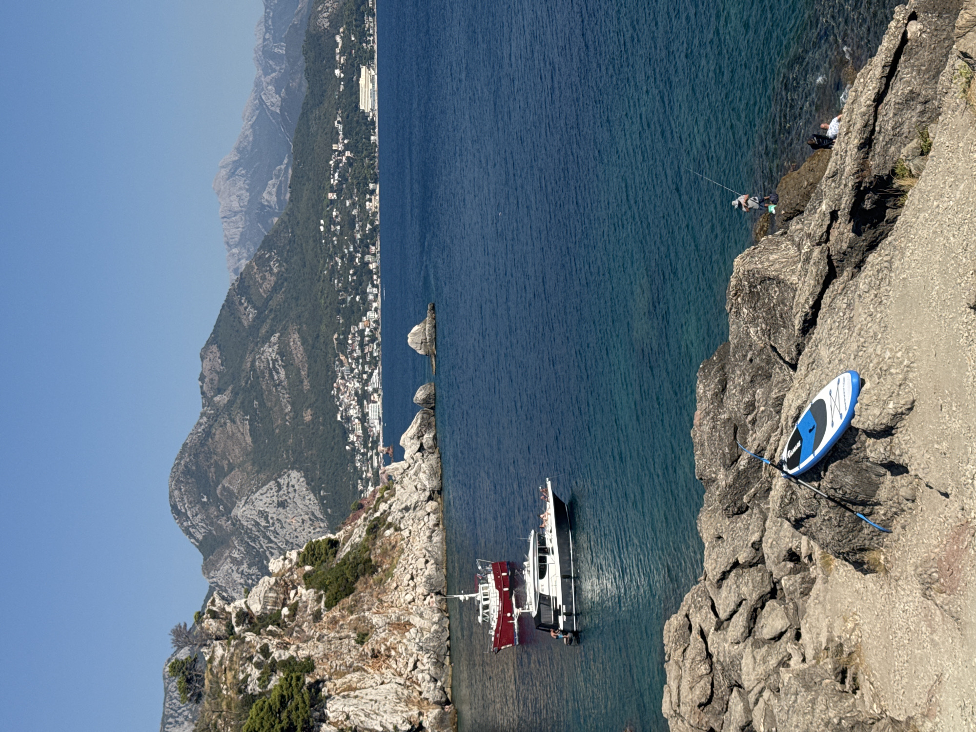 This is a medium shot, taken from a rocky outcrop, overlooking the Adriatic Sea towards Sutomore, Montenegro. The water is a deep, clear blue, with a rocky coastline in the foreground. Two boats are moored in the water: a small red and white tugboat and a larger, dark-hulled motor yacht. On the shore in the foreground, a blue and white paddleboard rests on the rocks. Further out in the sea, a lone figure stands on a rock, fishing. In the background, a densely populated coastal town with white buildings climbs up a forested hillside, with rugged mountains rising behind it under a clear blue sky.