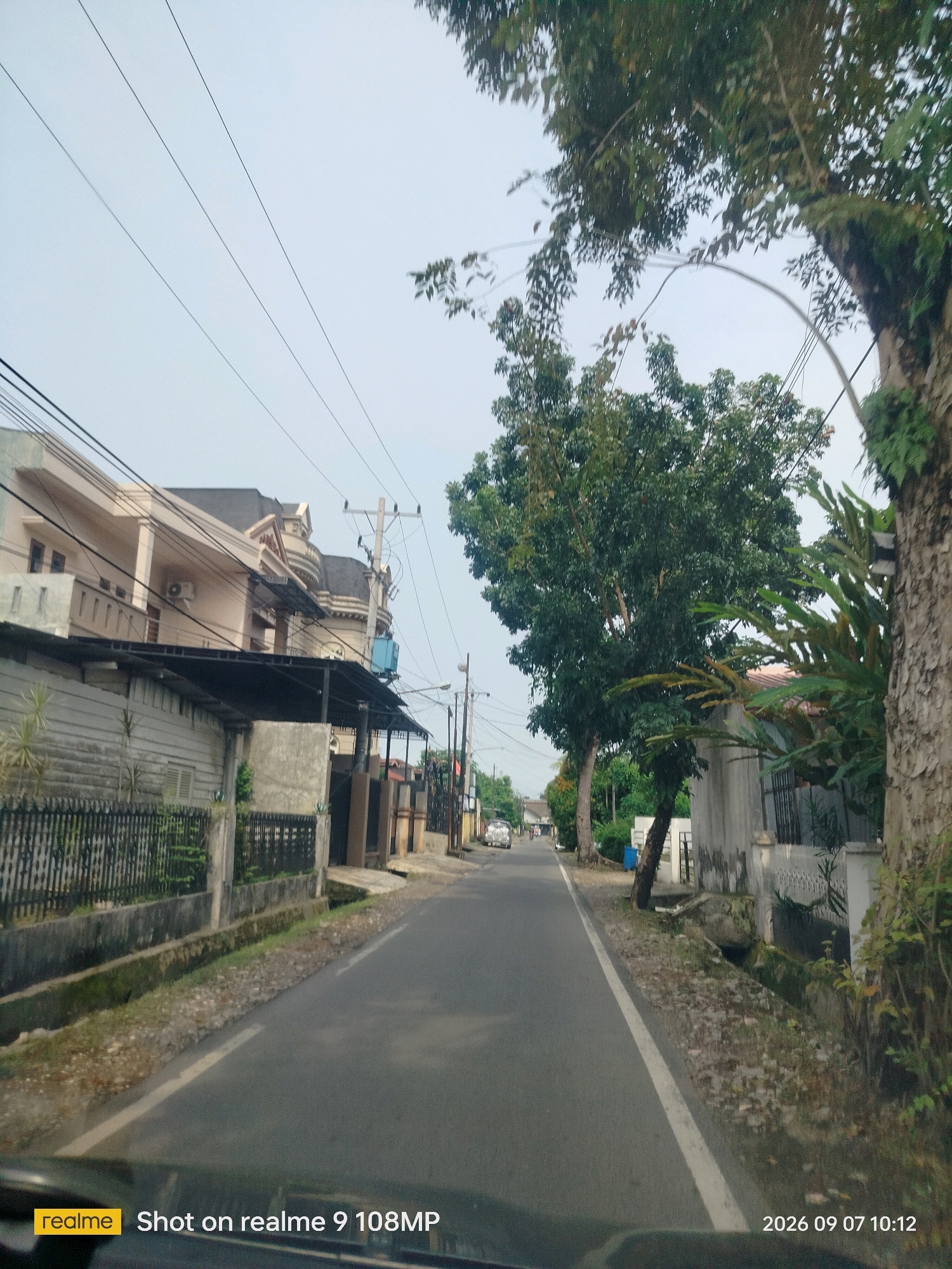A low-angle, wide shot captures a street scene in the city of Medan, Indonesia, on a clear day. The road, paved with asphalt, runs through a residential area lined with houses and trees. On the left side of the road, a modern, multi-story house with beige walls and dark grey accents stands behind a black metal fence. A black awning extends from a lower structure in front of the house, supported by thin poles. Electrical wires crisscross overhead, strung between utility poles.
To the right of the road, a large tree with a textured, greyish-brown trunk dominates the foreground. Lush green foliage surrounds its trunk, with a few branches extending towards the road. Behind this tree, more trees and vegetation are visible, creating a natural barrier between the street and other residences. Houses with lighter colored walls and tiled roofs are interspersed with the greenery further down the street. A small, light-colored car is visible in the distance on the left side of the road.
The ground along the sides of the road consists of gravel and dirt, with some areas of moss and grass. A narrow drainage ditch runs parallel to the road in several sections. The sky is a pale, hazy blue, suggesting an overcast or early morning/late afternoon setting. A watermark in the bottom left corner displays "realme Shot on realme 9 108MP", and a timestamp in the bottom right corner reads "2026 09 07 10:12".