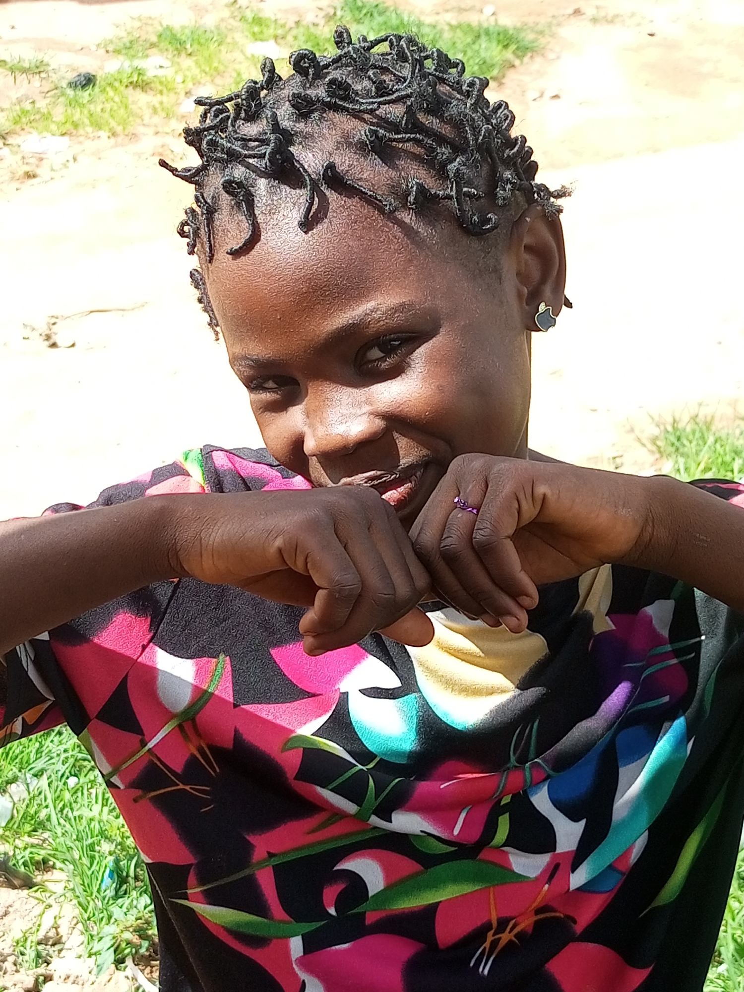 A medium-close shot captures a young Black girl with dark skin and braided hair in an outdoor setting, possibly Tempure, Nigeria. She is looking directly at the camera with a shy smile, her hands clasped in front of her face, partially obscuring her mouth. A small, dark earring with a silver outline is visible in her left ear, and a thin purple ring adorns a finger on her right hand. She is wearing a black t-shirt with a vibrant, abstract floral pattern in shades of pink, yellow, teal, and white. The background is softly blurred, revealing a sandy or dirt path with patches of green grass and some out-of-focus vegetation, suggesting a casual, natural environment. The lighting is bright, indicating daylight conditions.