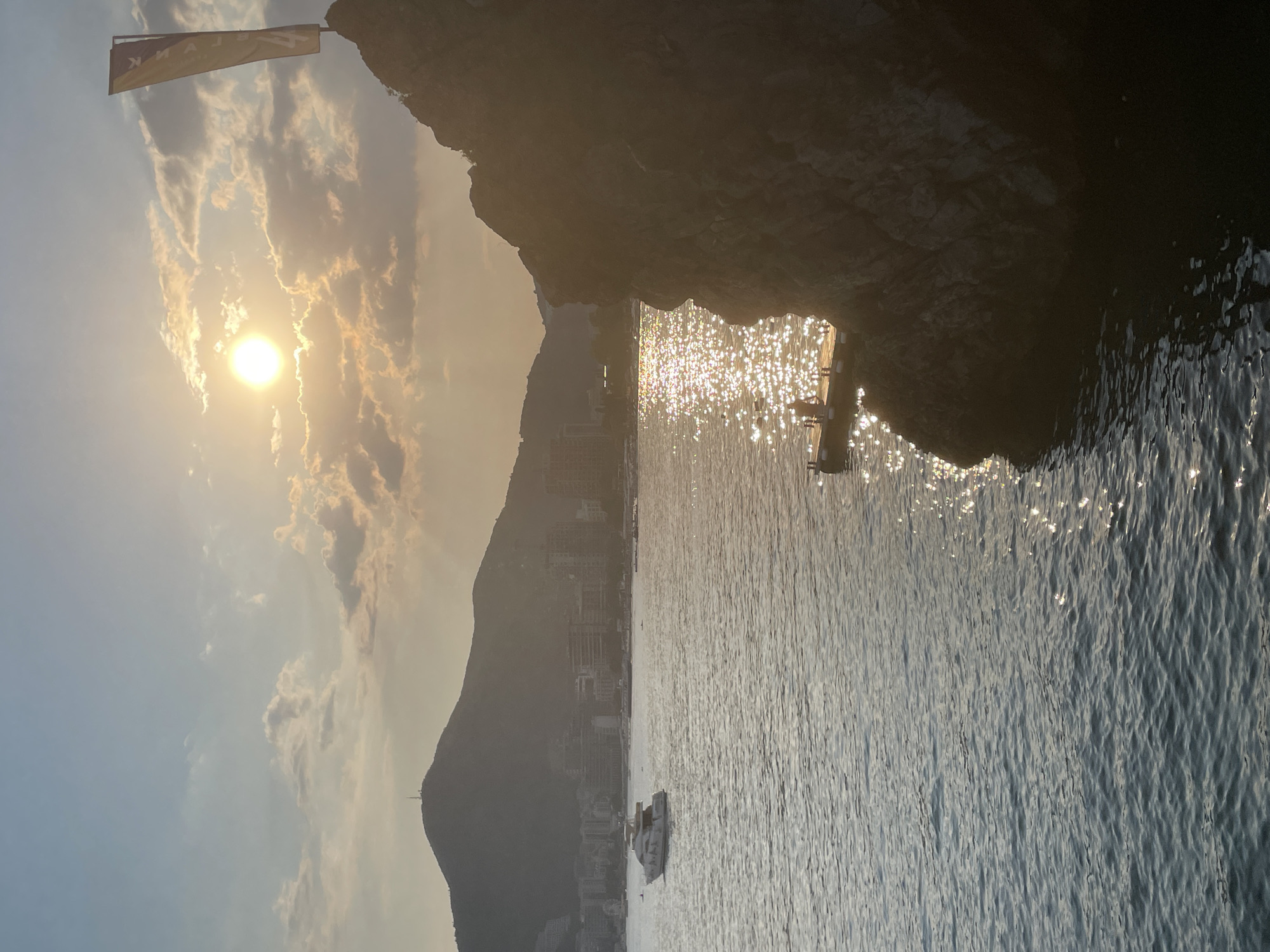 The image depicts a sunset over the Adriatic Sea in Budva, Montenegro. In the foreground, a rocky outcrop juts into the water from the right side of the frame. The surface of the sea is calm, with a shimmering reflection of the setting sun. A small wooden pier extends from the base of the rocky outcrop, with a single individual visible standing on it. In the middle ground, a white yacht is sailing on the water, and a distant shoreline is lined with buildings and hills. The sky is partly cloudy, with the sun positioned in the upper right quadrant of the image, casting a warm, golden light. A flag, bearing yellow and blue markings, is planted atop the rocky outcrop. The overall scene conveys a peaceful, serene atmosphere typical of coastal sunsets.