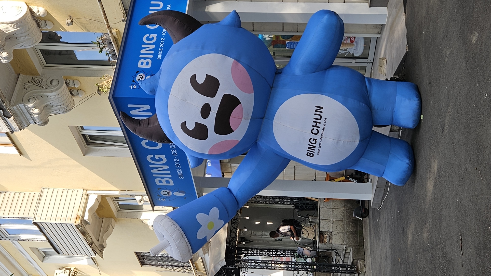 An inflatable blue mascot, resembling a cartoon character with brown horns and a large white circle on its chest with the text "BING CHUN SINCE 2012 - ICE CREAM & TEA," stands on a paved surface outside a building. The mascot is holding a large white cup with a blue floral design in its right hand. The building behind the mascot has a blue awning with the text "BING CHUN" and "SINCE 2012 - ICE CREAM & TEA." The building facade is yellow, with windows and architectural details. To the left of the mascot, there is a black metal gate and a person sitting on a bench. A scooter is parked near the entrance of the building. The scene appears to be on a street in Almaty, Kazakhstan.