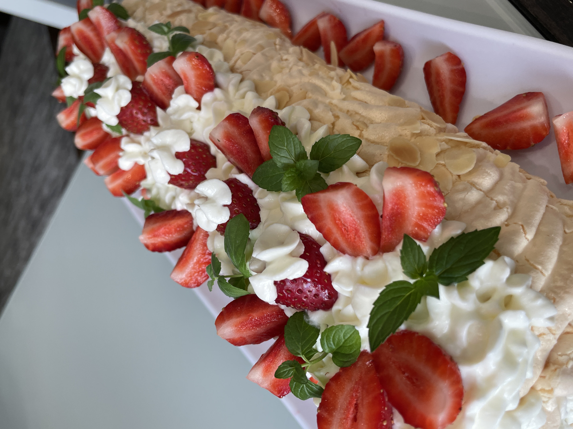 The image displays a close-up, eye-level view of a strawberry and cream roulade presented on a long, white serving dish. The roulade is topped with whipped cream, numerous sliced strawberries, whole strawberries, and fresh mint leaves. Sliced almonds are visible on the exterior of the meringue base. The background is out of focus, suggesting an indoor or sheltered outdoor setting. There are no people or discernible actions depicted. The image does not contain any information that would suggest a location in Saint Petersburg, Russia.