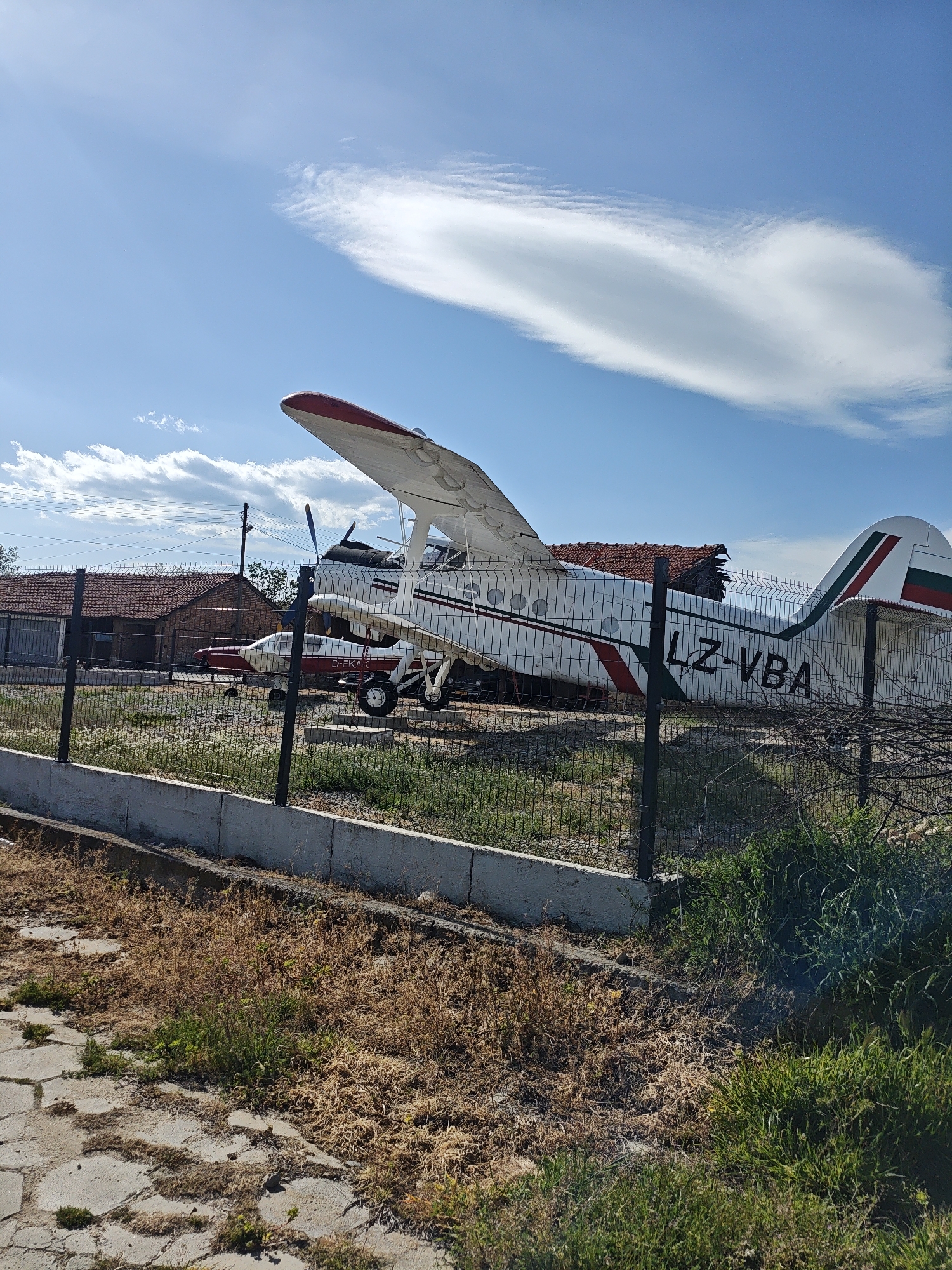 A medium shot shows two small aircraft behind a chain-link fence, with a tiled roof structure visible behind the airplanes. The larger aircraft, closer to the viewer, is white with red and green stripes, and the registration number LZ-VBA is visible on its tail. The second aircraft, behind and to the left of the first, is smaller and has a similar color scheme. The fence is a black chain-link style, supported by dark metal posts, and a short concrete wall runs along the base of the fence. The foreground shows a mixture of dry, brown vegetation and patches of green grass. A cracked concrete surface is visible in the lower-left corner. The sky is blue with scattered white clouds. The scene appears to be a display area or storage for decommissioned or vintage aircraft in the city of Unknown, Unknown.