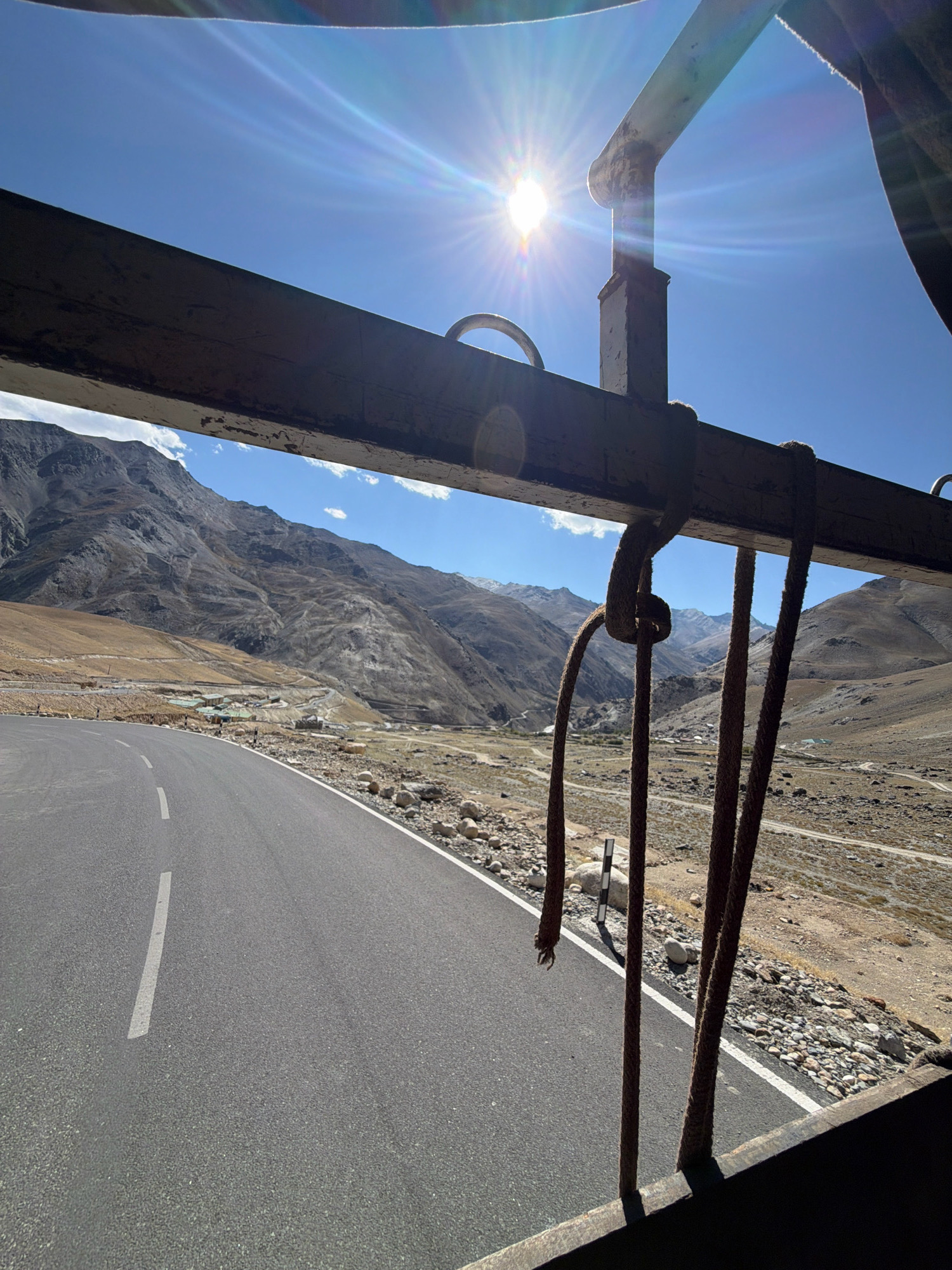 The image depicts a scenic mountain road in a rural, likely high-altitude location in India. A paved road curves to the left, bordered by rocky terrain and sparse vegetation. In the background, large, arid mountains dominate the landscape, with some snow visible on their peaks. A small village with clusters of buildings is nestled in a valley between the mountains. The sky is a clear, bright blue with a strong sun, casting lens flares and creating high contrast. In the foreground, several thick, brown ropes hang from an unseen structure, obscuring parts of the view and suggesting the perspective is from a vehicle or elevated platform. The overall impression is one of a remote, mountainous region under a clear, sunny sky.
