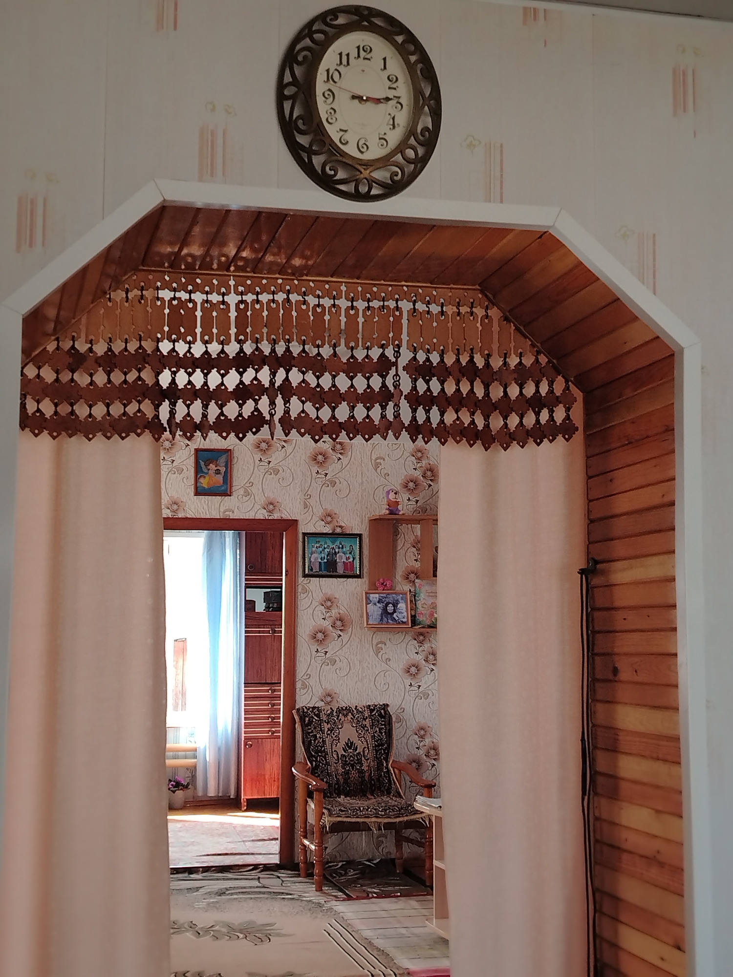 The image shows an interior view through an archway in what appears to be a residence in Яган, Russia. The archway is trimmed with light-colored wood and has a decorative wooden valance hanging within the opening. A clock hangs on the wall above the archway. A sheer curtain drapes on either side of the archway. The room visible through the archway has floral wallpaper and contains a chair with patterned upholstery, a small shelf unit holding various items, and framed pictures on the wall. A doorway leading into another room is visible, with a wooden cabinet and a window covered by a light blue curtain. The floor is covered with a rug or carpet.