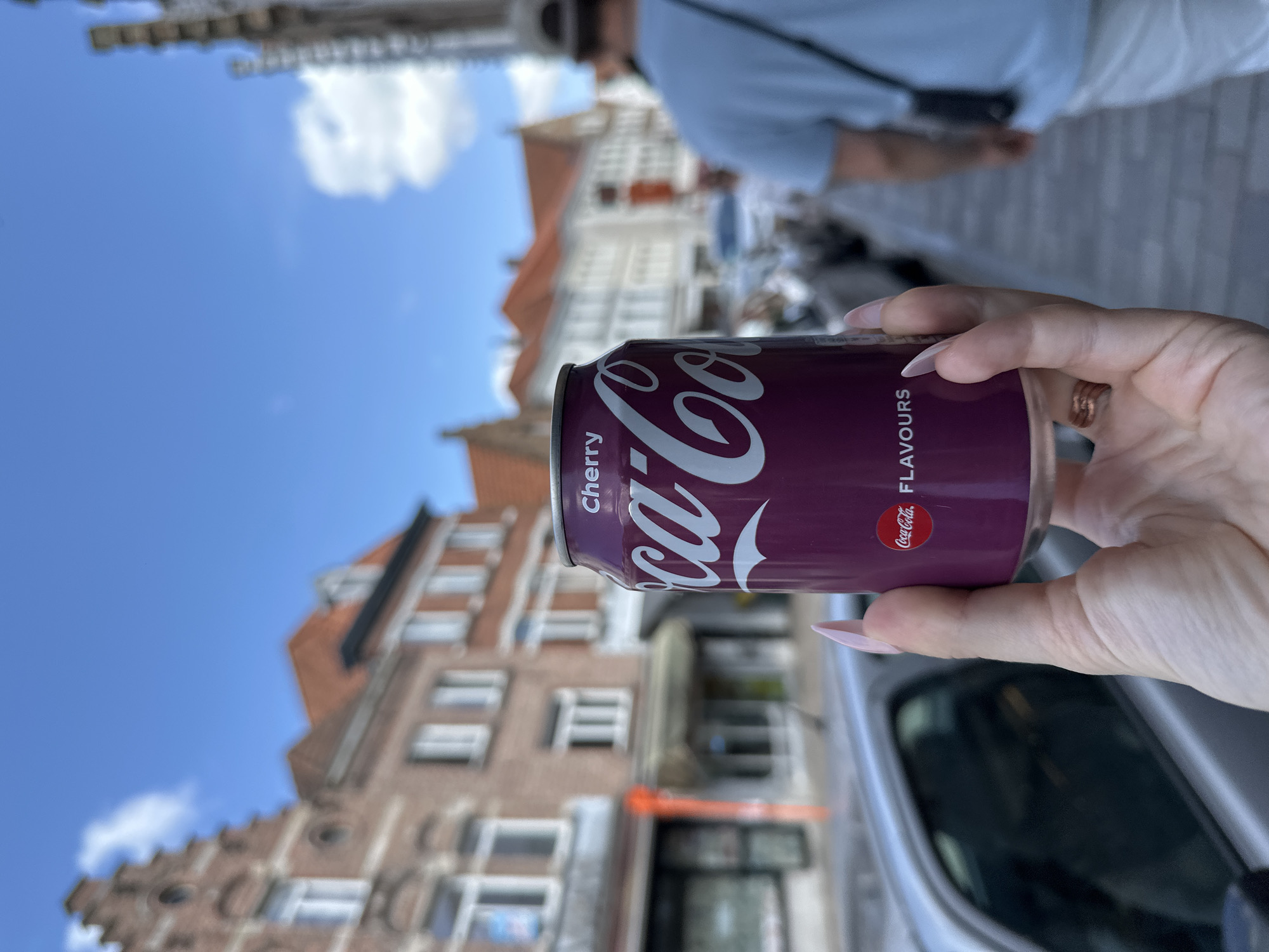 A person's hand, adorned with light pink manicured nails and a rose gold ring, holds a can of Coca-Cola Cherry Flavours. The can is a deep purple color with white lettering. In the background, a street scene unfolds in Bruges, Belgium. Buildings with traditional Flemish architecture, characterized by stepped gables and red-tiled roofs, line the street. The sky is blue with a few scattered white clouds. Cars are parked along the street and a person wearing a light blue t-shirt and white pants walks away from the camera. The overall impression is a snapshot of a casual moment in a historic European city.