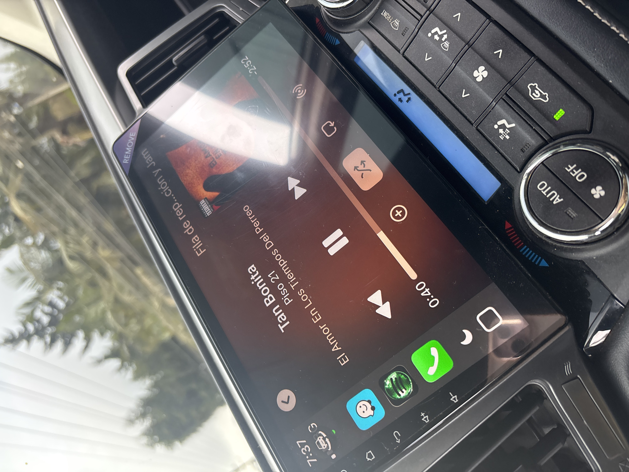 The image shows the dashboard of a car. A screen displaying music is in the center, with the song "Tan Bonita" by Piso 21 playing from the album "El Amor En Los Tiempos Del Perreo." The time on the screen reads 7:37. To the right of the screen, climate control buttons are visible. The car's location is inferred to be in Guarne, Colombia, based on the displayed song information. The scene contains no people.