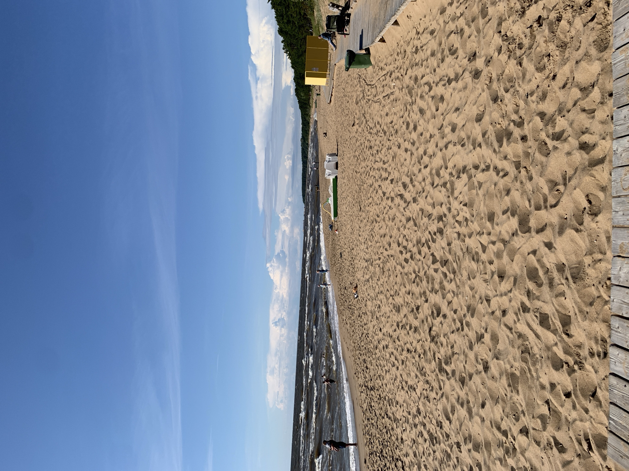 The image shows a sandy beach in Saulkrasti, Latvia. The sky is blue with scattered white clouds. The sea is choppy with waves. Several people are visible on the beach and in the water. There are some beach chairs and a yellow structure that appears to be a changing room or lifeguard station. A wooden boardwalk runs along the edge of the beach. The sand is rippled, indicating it has been recently disturbed by wind or footsteps. The overall impression is a sunny day at the beach.