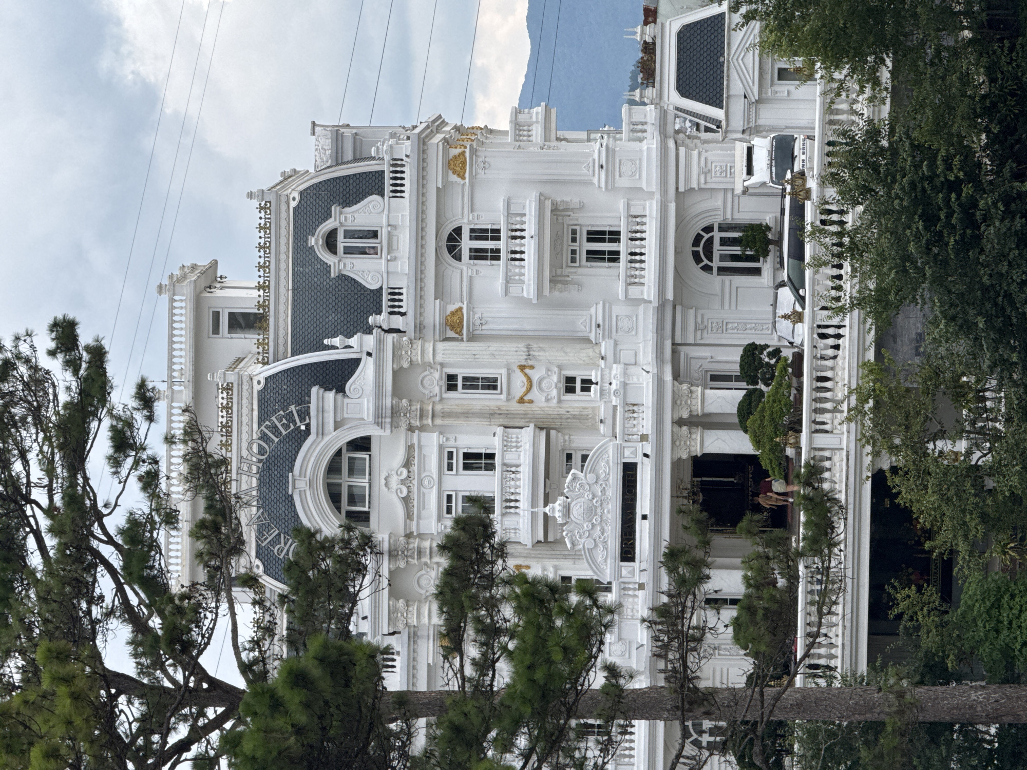 The image shows the exterior of a large, ornate white hotel with a dark gray roof, nestled amongst lush green trees. The hotel features multiple levels with balconies, arched windows, and decorative gold accents. The name "DREAM HOTEL" is prominently displayed on the front facade, visible on the roofline and above the main entrance. In front of the hotel, a white railing overlooks a lower level which appears to be a restaurant with signage. Several cars are parked on the hotel's grounds, including a white truck with text that is partially visible. Two people are seen walking on the upper balcony near the entrance. The background reveals distant mountains under a cloudy sky. The hotel's elaborate architectural style suggests it is a notable landmark within Da Lat, known for its French colonial influences and picturesque landscapes.