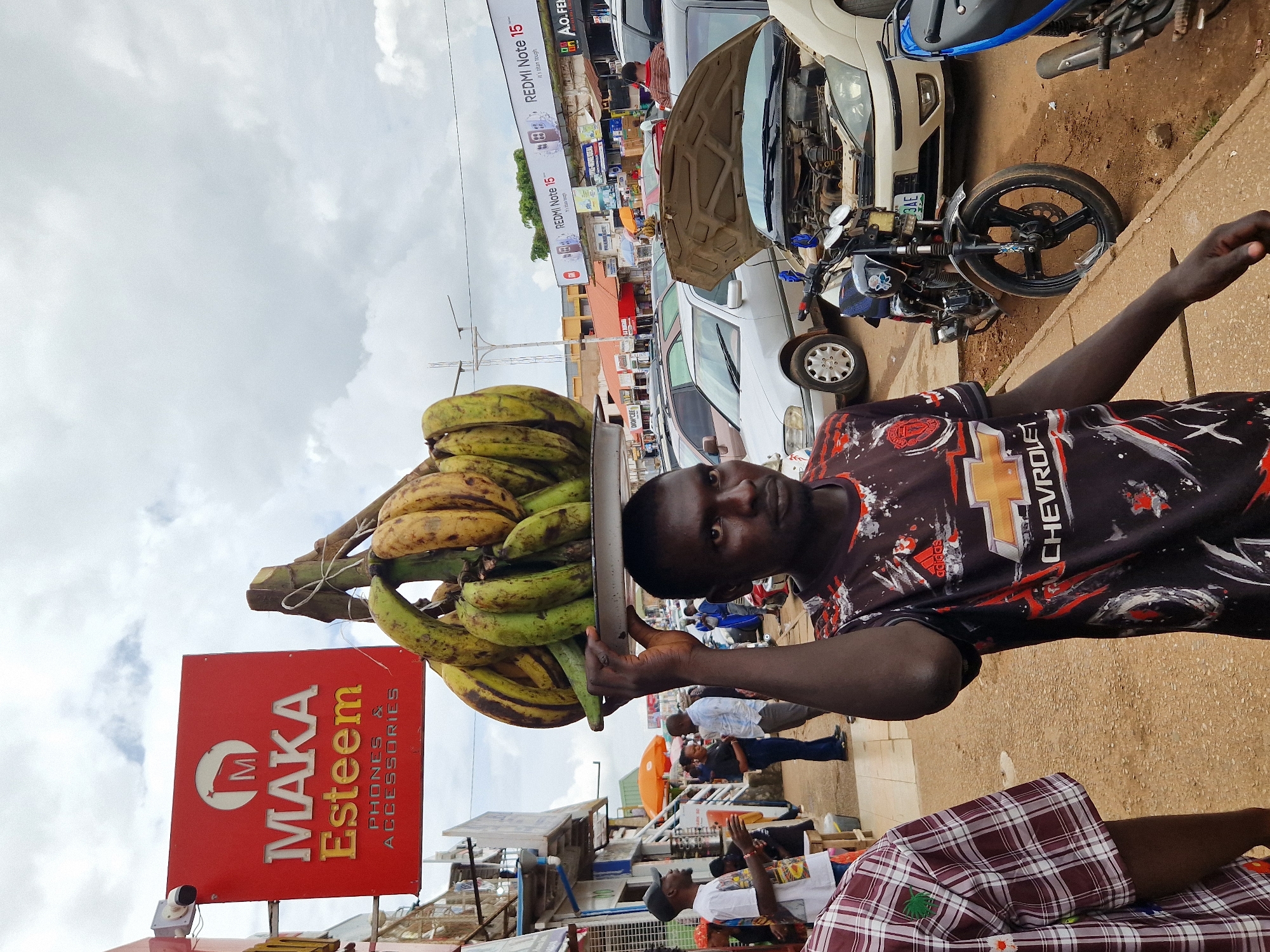 A dark-skinned man is depicted from the chest up, balancing a tray of ripe bananas on his head. He wears a black t-shirt with a red and white abstract pattern and a Chevrolet logo. The background shows a busy street scene. To the left, a red sign with a white 'M' logo reads "MAKA Esteem PHONES & ACCESSORIES". Further back, a street lined with shops and parked cars is visible under an overcast sky. A white sedan with its hood open is parked in the middle ground, alongside motorcycles. The ground is a mix of dirt and pavement.