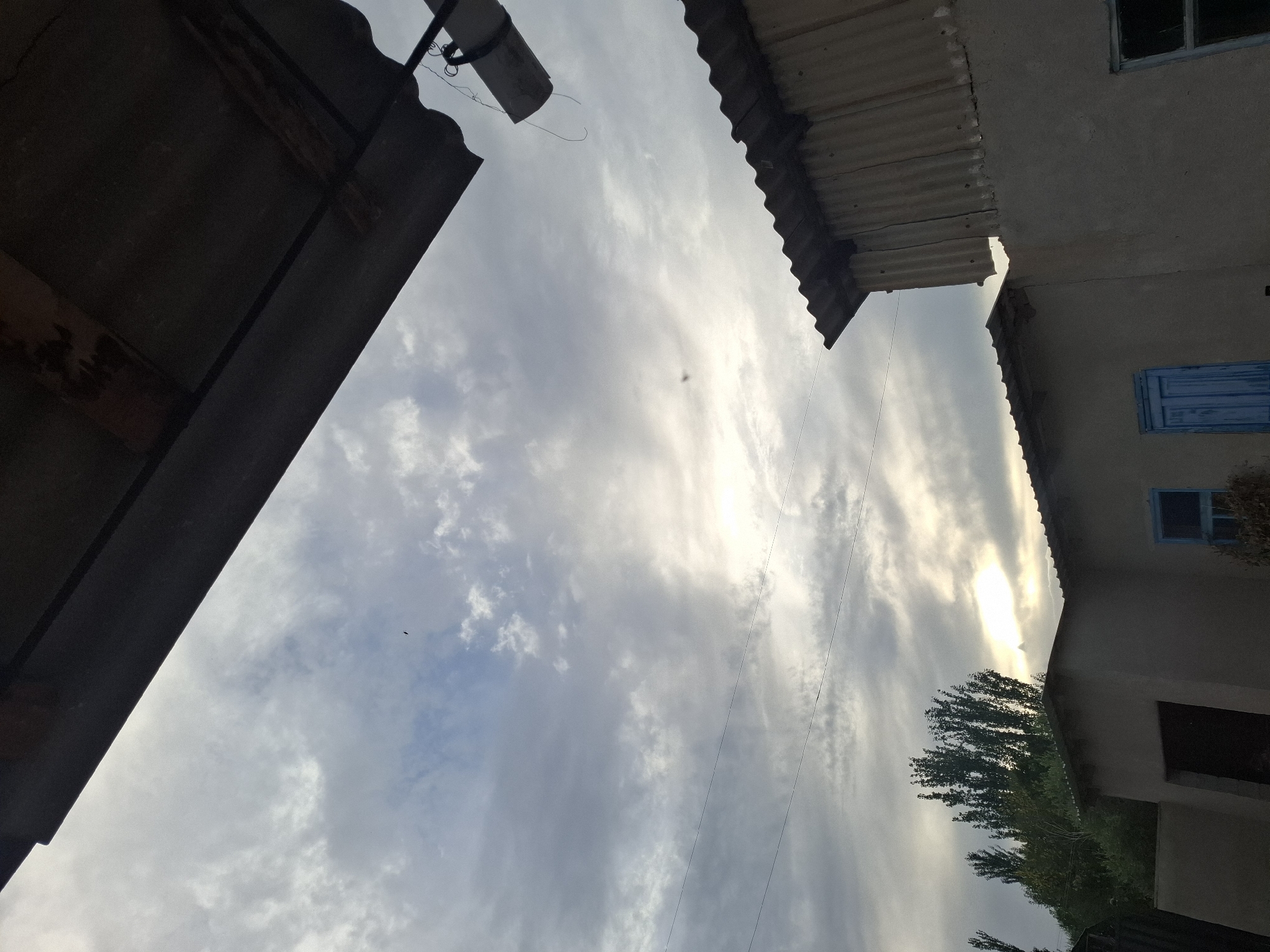 The image depicts an overcast sky with scattered clouds and a muted sun. In the foreground, several weathered, corrugated metal roofs are visible, attached to low-lying structures that appear to be part of a residential or utility area. One building has a blue wooden door and a window. To the left, a cluster of tall, dark green trees stands out against the sky. Two thin, dark lines, likely power or telephone lines, stretch horizontally across the sky. In the upper part of the image, a dark, angular overhang, also made of corrugated material, dominates the scene. A cylindrical object, possibly a light fixture or a ventilation pipe, hangs from the overhang. The overall impression is of a utilitarian, possibly rural or semi-urban, environment in the Jeti-Ögüz region of Kyrgyzstan, under a dull sky. No people are visible in the image.