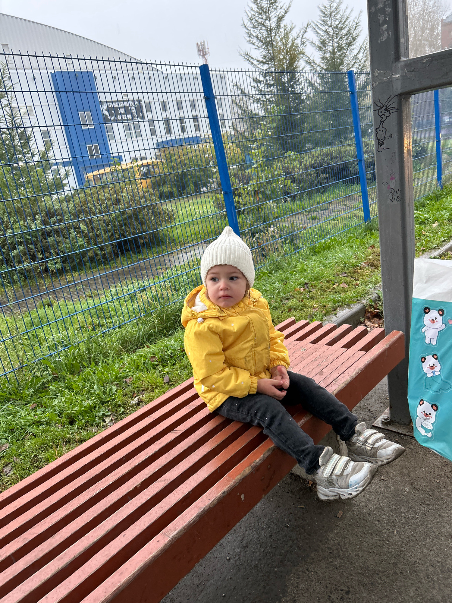 A young child, dressed in a yellow jacket and a white beanie, sits on a reddish-brown slatted park bench. The child is looking to the right with a neutral expression. The bench is situated outdoors, with green grass and some fallen leaves on the ground. In the background, a blue chain-link fence separates the seating area from a grassy path and trees. Behind the fence, a building with white siding and blue accents is visible, along with a yellow school bus. A gray utility pole with graffiti is to the right of the bench, and a light blue bag with panda graphics is hanging from it. The scene appears to be in an outdoor public space, possibly near a school or park, in Tomsk, Russia, as indicated by the presence of a school bus and the architectural style of the building. The weather seems overcast and damp.