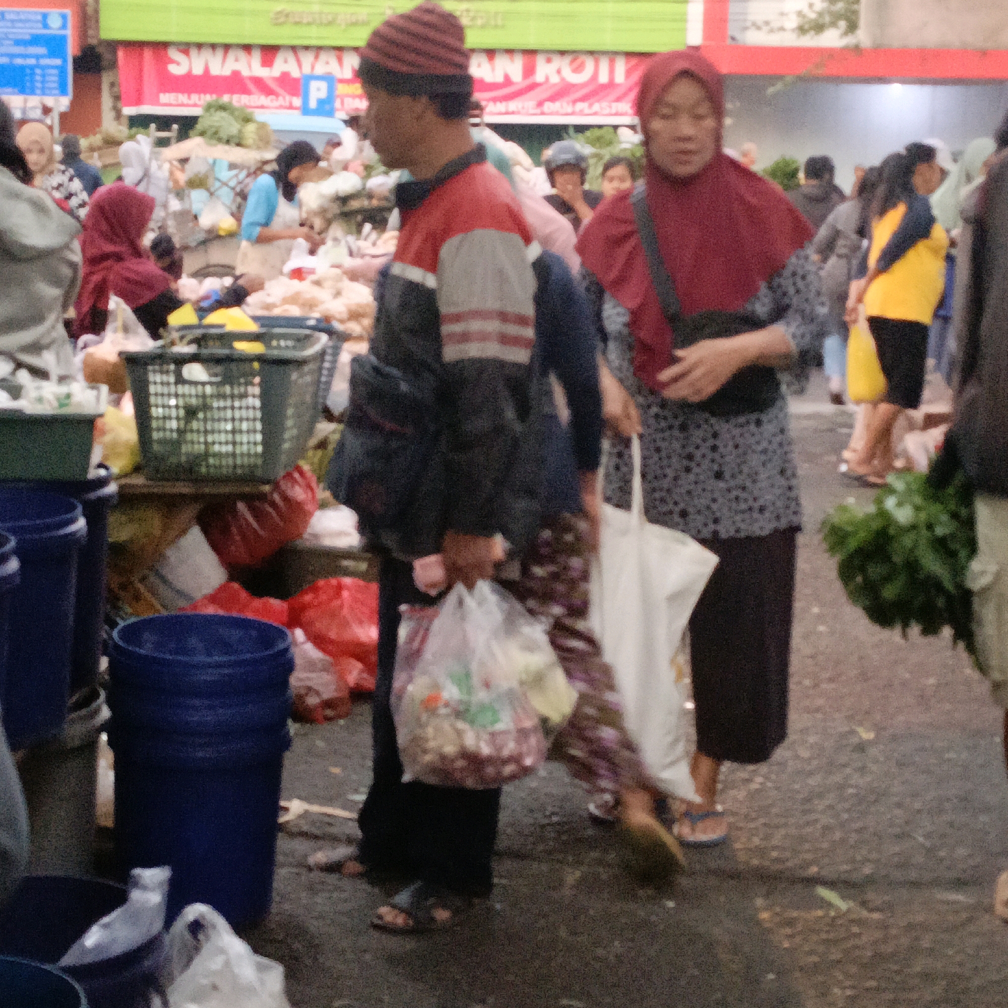 The image depicts a bustling outdoor market scene in Salatiga, Indonesia. In the foreground, a person wearing a red and gray jacket and a striped beanie walks to the left, carrying a black bag. To their right, a person in a red hijab and patterned shirt holds a white tote bag and walks away from the camera. Behind them, another person in a red hijab and patterned shirt is visible. Further back, a woman in a yellow and blue top stands near a pile of green vegetables, and other shoppers are seen amidst stalls and goods. Various baskets, buckets, and plastic bags filled with items are scattered throughout the market. The overall environment suggests a typical day at a local Indonesian market.