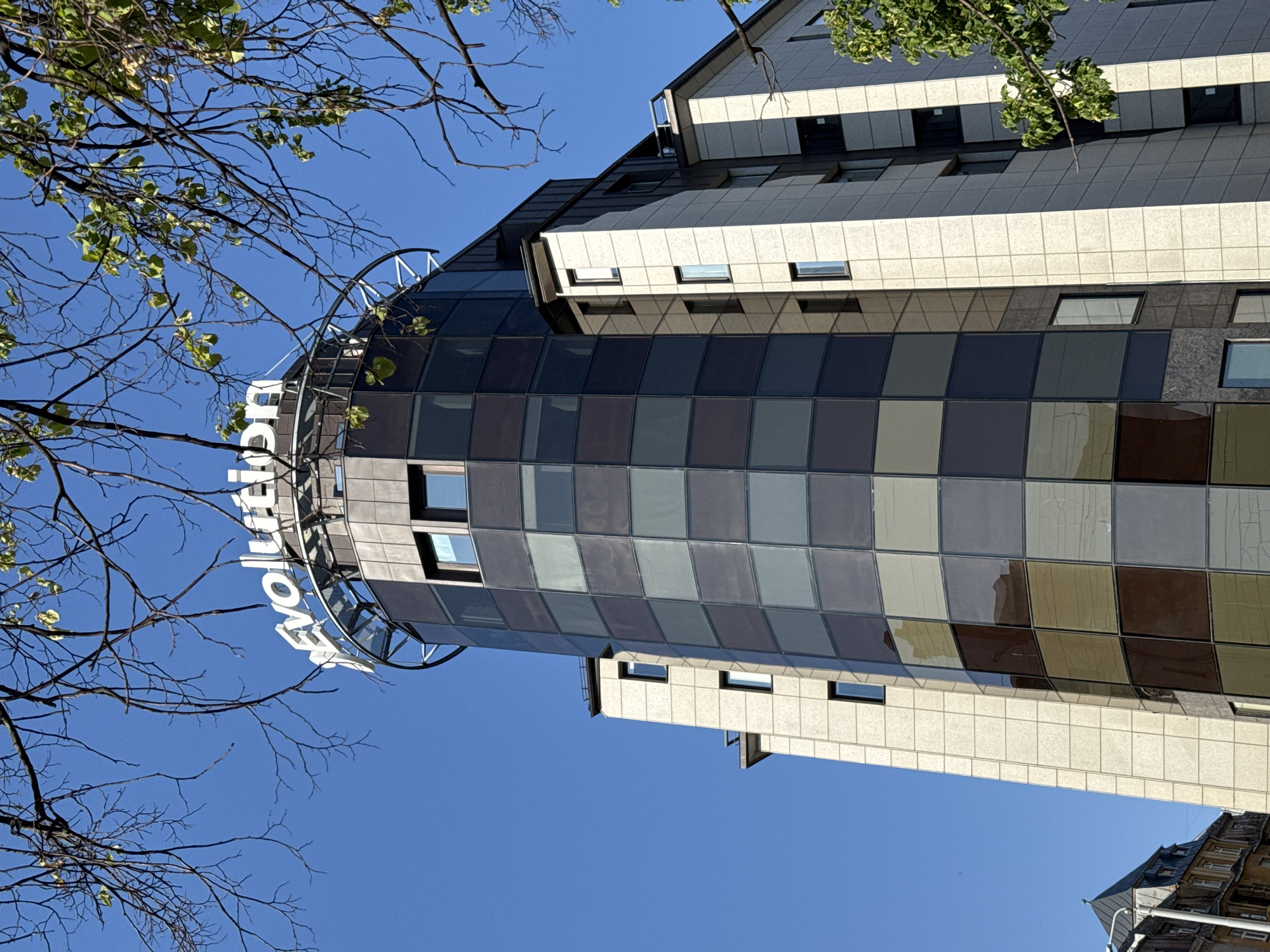 An upward-angled outdoor shot captures a modern building against a clear, vibrant blue sky. The main subject is a multi-story structure, likely commercial or a hotel, showcasing a distinctive architectural design. Its prominent upper section features a curved facade composed of a grid of glass or reflective panels, exhibiting various shades of grey, blue, brown, and beige, creating a mosaic-like pattern. Visible windows are interspersed throughout this section.
Adjacent to and below the curved facade, the building incorporates sections with light-colored, possibly stone or concrete, cladding, and dark-tiled, sloped roofs, suggesting an attic or mansard level. Numerous windows are visible across these different parts of the building.
In the foreground, bare tree branches with a scattering of fresh green leaves partially frame the sky and the building, adding a natural element to the urban scene. On the curved glass section of the building, large white letters spell out a portion of the word "Evolution," indicating the name of the establishment. A small fragment of another, older-style building with a gabled roof is visible in the bottom right corner, suggesting an integrated urban setting.
The scene is brightly illuminated by natural daylight, indicating a clear and sunny day. No specific activity or event is taking place; it's a static architectural view. The visible text and provided context confirm the location as Riga, Latvia.