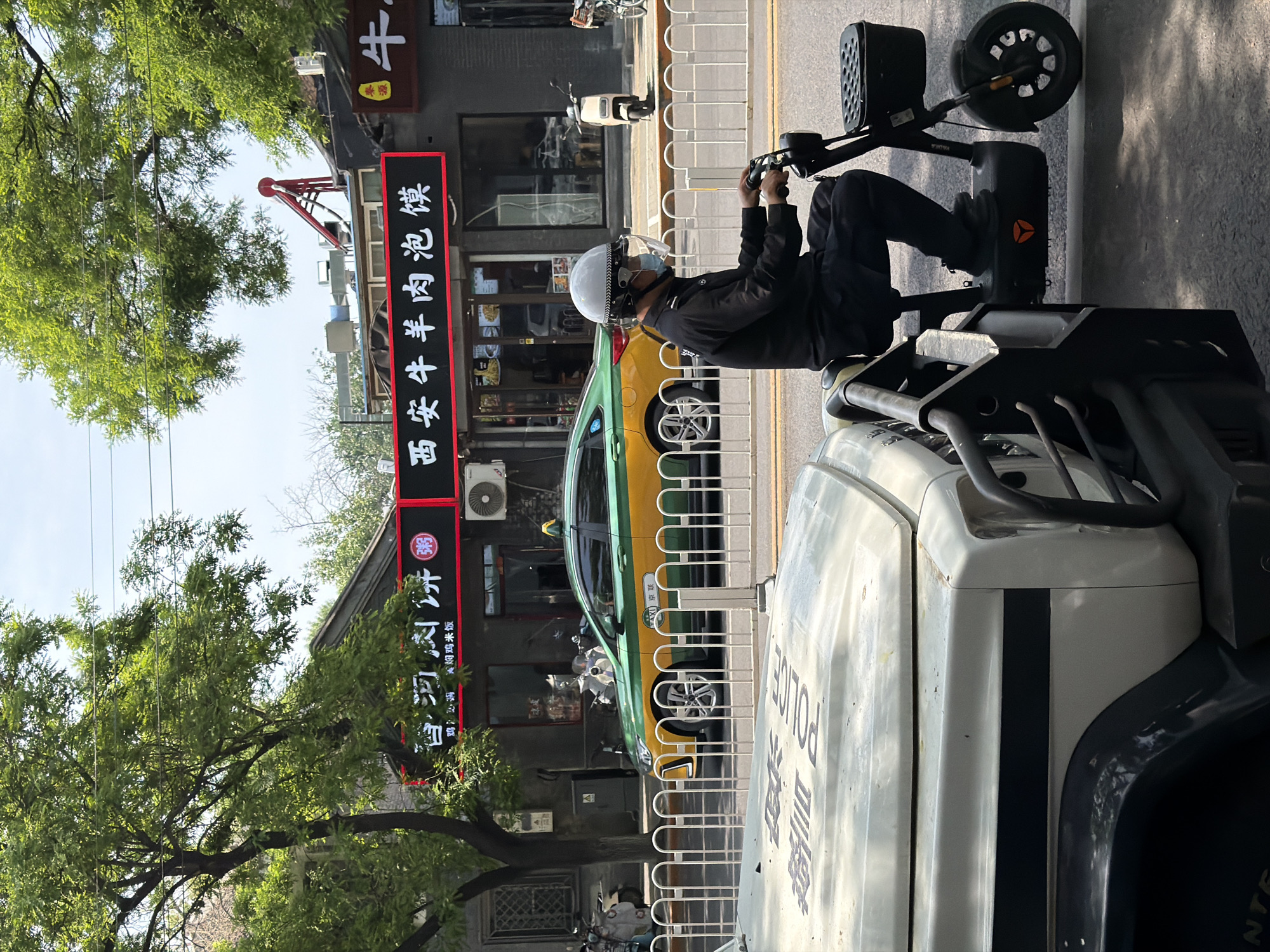 A daytime outdoor shot shows a man on a three-wheeled electric scooter next to a Beijing police vehicle in an unidentified area of Dongcheng District, China. The man, wearing a white helmet and a black jacket, sits on the scooter, his hands on the handlebars. A yellow and green taxi sits parked behind a fence. Behind the taxi is a two-story building with a storefront sign in Chinese characters advertising food. The area is likely commercial with sidewalks and pedestrian barriers visible. A tree is visible in the foreground on the left side of the image. The police vehicle is marked with the word "POLICE."