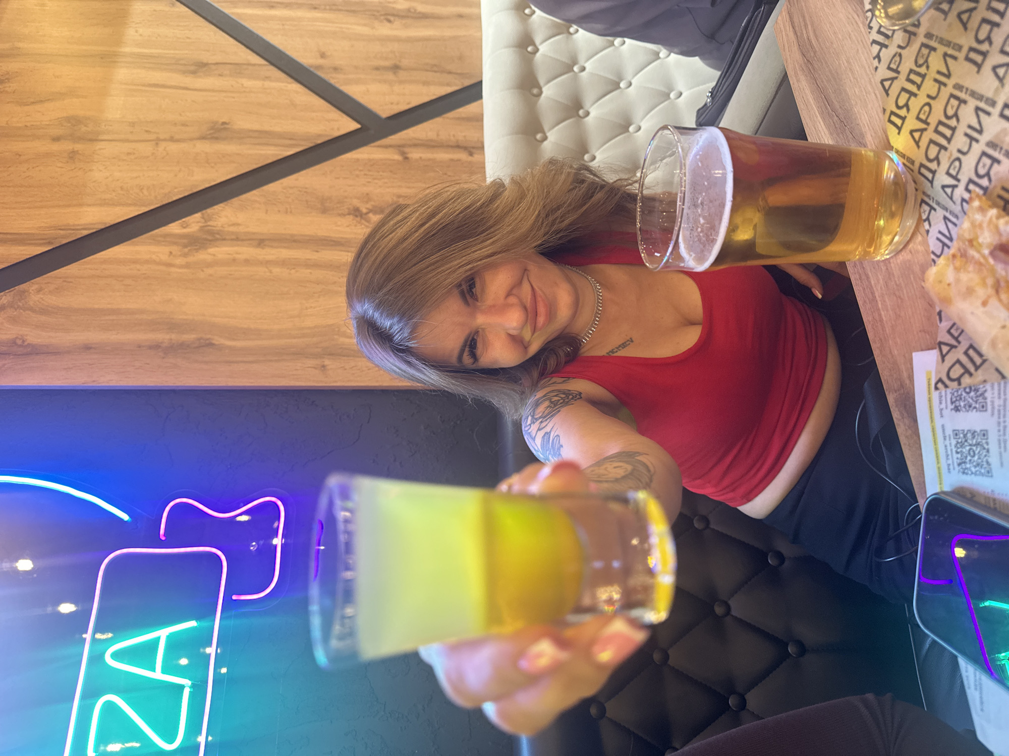 The image shows a person indoors, likely at "Uncle Archi" bar in Perm, Russia, based on the signage visible. They are holding a shot glass filled with a yellow liquid and a beer glass with a golden liquid. The person has light brown hair, a red top, and tattoos on their left arm. There is a table with wooden surface and a menu with QR codes on it, alongside a phone. A neon sign is visible in the background.