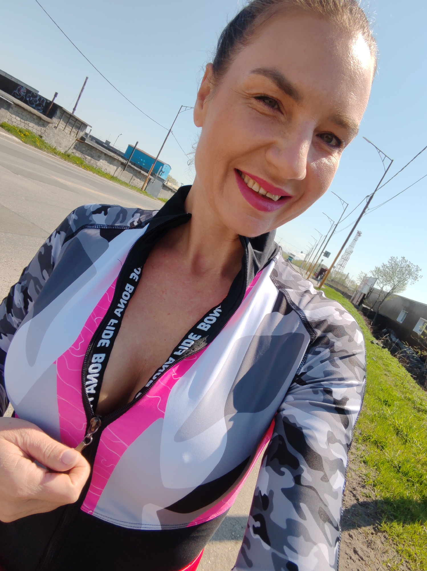 The image depicts a female individual in Petropavlovsk-Kamchatsky, Russia, captured from a low-angle perspective under clear daylight. The woman, appearing to be in mid-adulthood, is smiling broadly at the camera, wearing bright pink lipstick and with her hair pulled back. She is dressed in athletic wear, specifically a long-sleeved top featuring a grey, black, and white camouflage pattern, accented with a pink and white design across the chest. The front zipper of the top is partially open, revealing a pink internal band with the inverted text "BONA FIDE BONA FIDE" repeatedly printed. Her left hand is visible, holding the zipper pull. Portions of matching camouflage leggings are also visible.
The background shows an outdoor environment with an asphalt road or path extending to the left, bordered by green grass with visible yellow dandelions. Beyond the road, a low concrete-block wall with graffiti markings is present, followed by industrial-style buildings, one of which is blue. Streetlights are visible along the road. To the right, a grassy embankment and a dirt path are discernible, along with a small, largely leafless tree and a low-lying building structure. In the distant background, a tall, lattice-structured communication tower stands against a clear, light blue sky.