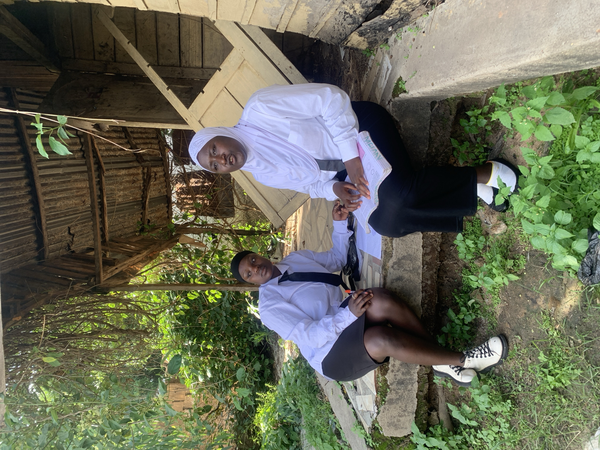 Two adult females are seated outdoors on a concrete retaining wall or ledge, engaged in an apparent discussion or study session. The scene is set in Jos, Nigeria, amidst a mix of natural vegetation and rustic structures.
The female on the left, with dark skin, wears a white long-sleeved button-up shirt, a black necktie, and a black knee-length skirt. Her dark hair is covered by a black cap or head wrap. She is seated with her legs bent and feet resting on the ground, wearing white and black lace-up shoes. Her right hand appears to hold a pen near an open notebook or pad and a small black handbag positioned between the two individuals on the concrete ledge. Her gaze is directed slightly past the viewer, maintaining a serious expression.
The female on the right, also with dark skin, wears a white long-sleeved button-up shirt, a black long skirt, and a white hijab covering her head and chest over a dark underscarf. She is seated with her legs slightly extended, wearing black slip-on shoes with white socks that have a yellow top band. Her hands rest on an open book with a pink and white cover. Her mouth is slightly open, suggesting she is speaking or in mid-sentence, and she appears to be addressing the other woman.
The immediate foreground consists of bare earth with scattered green ground cover and small plants. The background to the left is dominated by dense, lush green vegetation, including numerous hanging vines and trees, with sunlight filtering through. A reddish-brown structure is partially visible through the foliage. To the right, a dilapidated wooden structure, possibly an outbuilding or shed, features weathered wooden planks and a corrugated metal roof. A black plastic bag is visible on the ground near the base of this structure. The scene is illuminated by natural daylight.