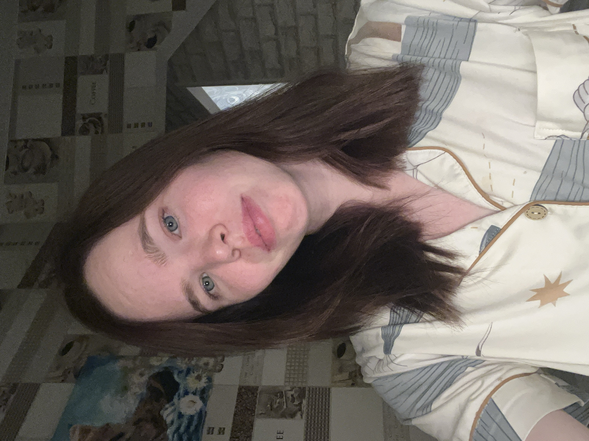 A woman with brown hair and blue eyes is lying down, looking directly at the camera. She is wearing a patterned pajama top with a button closure and a star design. The background consists of tiled walls, one section with a "Coffee" motif and another with a painting. The location is suggested as the city of Chaikovsky, Russia.