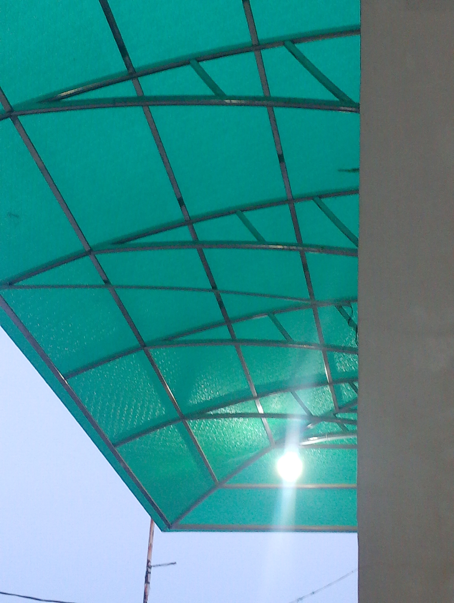 The image shows a close-up, upward-angled view of a translucent green roof structure, likely made of polycarbonate or a similar material, supported by a dark metal frame. The roof is composed of rectangular panels arranged in a grid pattern, with some diagonal supports. A bright light bulb is illuminated beneath the roof, causing a lens flare and casting a glow. To the right of the roof structure, a light gray, textured wall or pillar is visible. In the background, the sky is a pale blue. A thin pole or support structure extends upwards from below the frame on the left side, with some wires visible near the bottom of the image. The scene appears to be outdoors, possibly a covered walkway, patio, or an awning in an urban setting. Without further context or identifying markers, it is not possible to pinpoint the specific location within Batam, Indonesia.